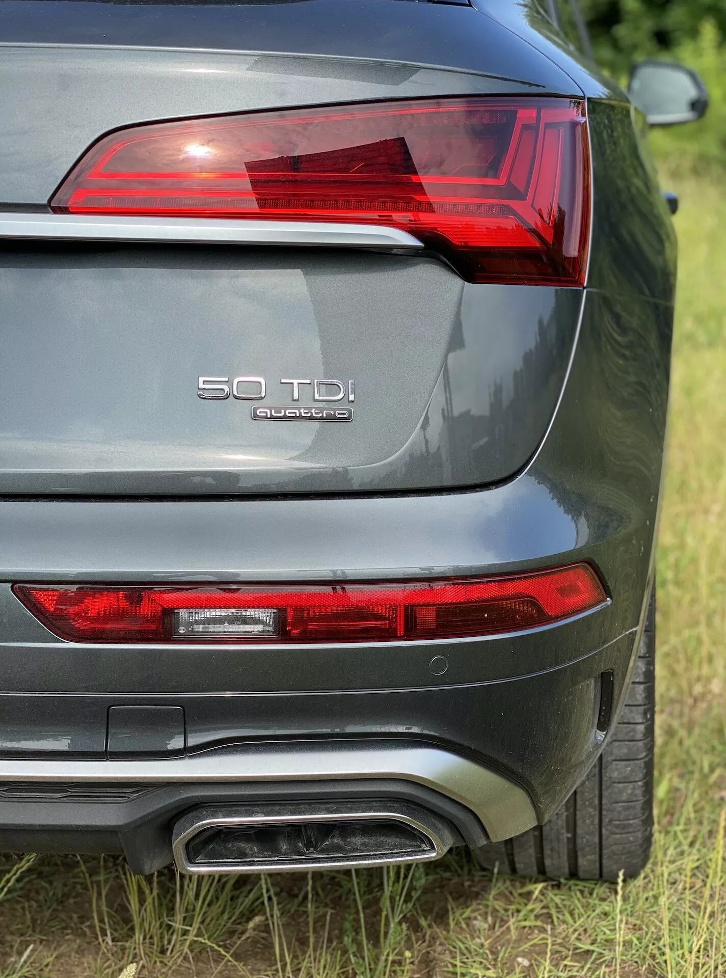 Vue rapprochée de la section arrière de l'Audi Q5 2020 Sportback depuis une perspective à trois quarts arrière, montrant le feu arrière gauche avec des éléments LED, la désignation du modèle '50 TDI' et 'quattro' sur le couvercle du coffre, un système d'échappement double, une peinture métallisée grise, et un capteur de stationnement arrière visible.