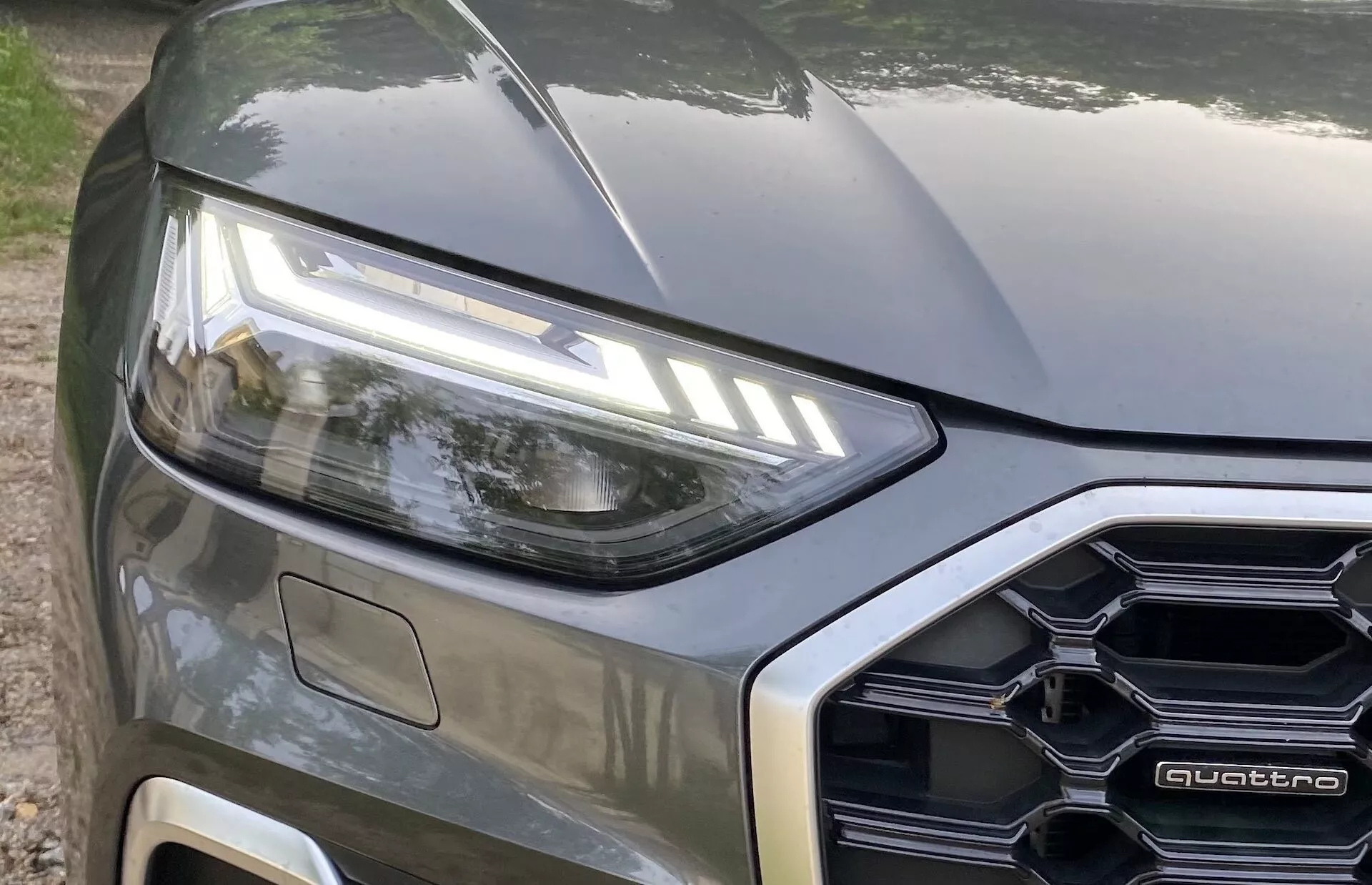 Gros plan du côté avant gauche d'une voiture moderne, montrant une vue partielle incluant le phare LED gauche, une partie du capot et une partie de la calandre avant de conception polygonale avec un badge 'quattro' visible. La prise de vue capture les éléments de design de la voiture et la technologie d'éclairage avec une finition de peinture métallique, prise d'une perspective de niveau légèrement inclinée vers le haut.