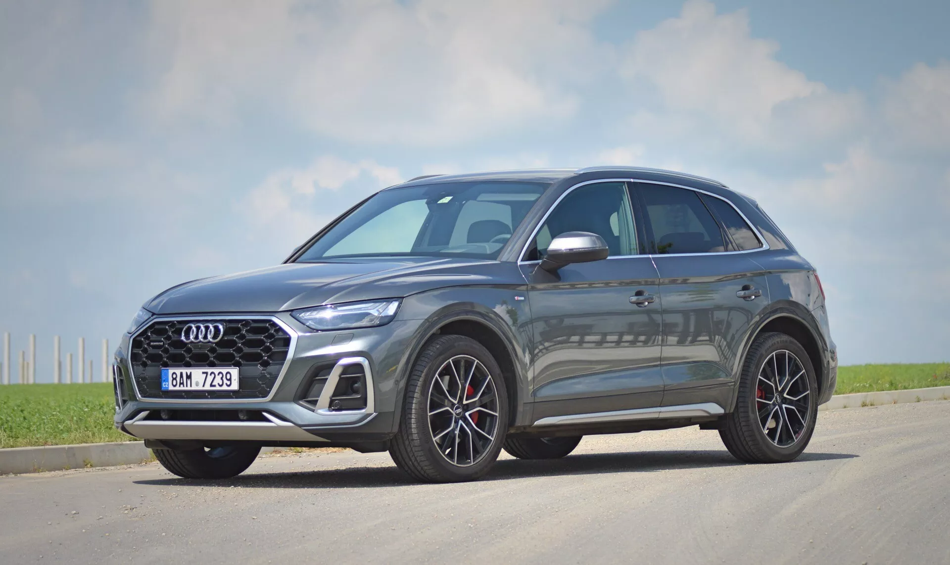 Image d’un crossover/SUV moderne, le modèle Audi Q5 Sportback 2020, stationné sur un tronçon de bitume. La voiture est présentée en vue de trois quarts avant, capturant à la fois le devant et le profil latéral. La grille proéminente avec un motif en croix noir brillant présente l'emblème du fabricant au centre. Les phares LED élégants et pointus et les roues en alliage à rayons multiples rehaussent l'esthétique haut de gamme. La peinture métallique reflète la lumière du soleil, indiquant une finition de haute qualité. Le profil latéral montre des lignes de carrosserie aérodynamiques et des rétroviseurs extérieurs avec clignotants intégrés. Les fenêtres ont une garniture chromée contrastant avec les vitres teintées, suggérant éventuellement un vitrage intimité. La voiture est immobile, positionnée légèrement à droite de l'axe central de la caméra avec des ombres suggérant qu'il est soit matin soit après-midi, sous un ciel clair avec quelques nuages dans un environnement rural ou semi-rural.