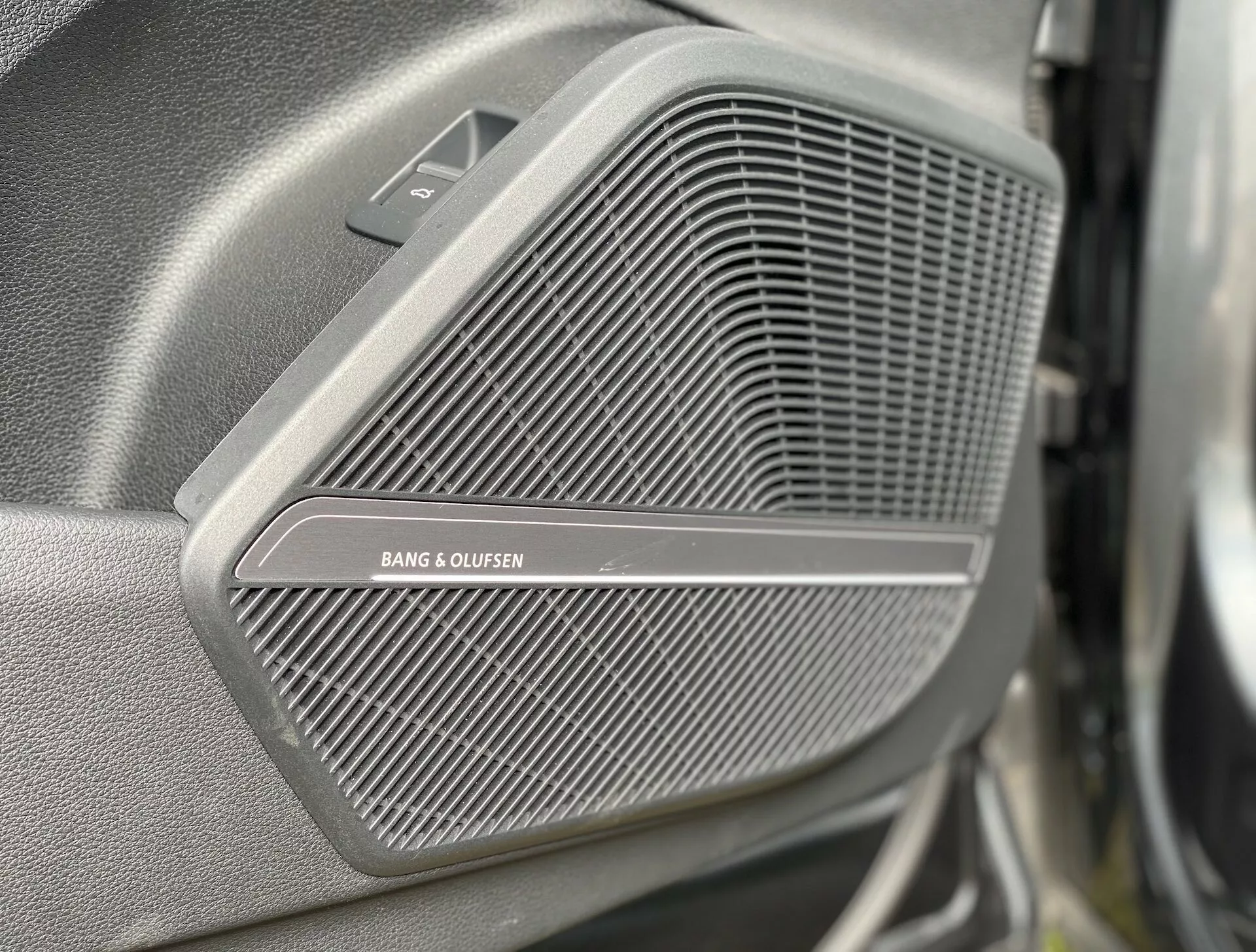 Gros plan sur la grille du haut-parleur premium BANG & OLUFSEN sur le panneau de porte de l'Audi Q5 Sportback 2020. Le motif en treillis fin de la grille est net, tandis que l'arrière-plan et le premier plan sont flous. Au-dessus de la grille, une petite section triangulaire, visible sous un angle légèrement latéral et avant, pourrait abriter un tweeter. L'image met en avant la qualité luxueuse du système audio du véhicule.
