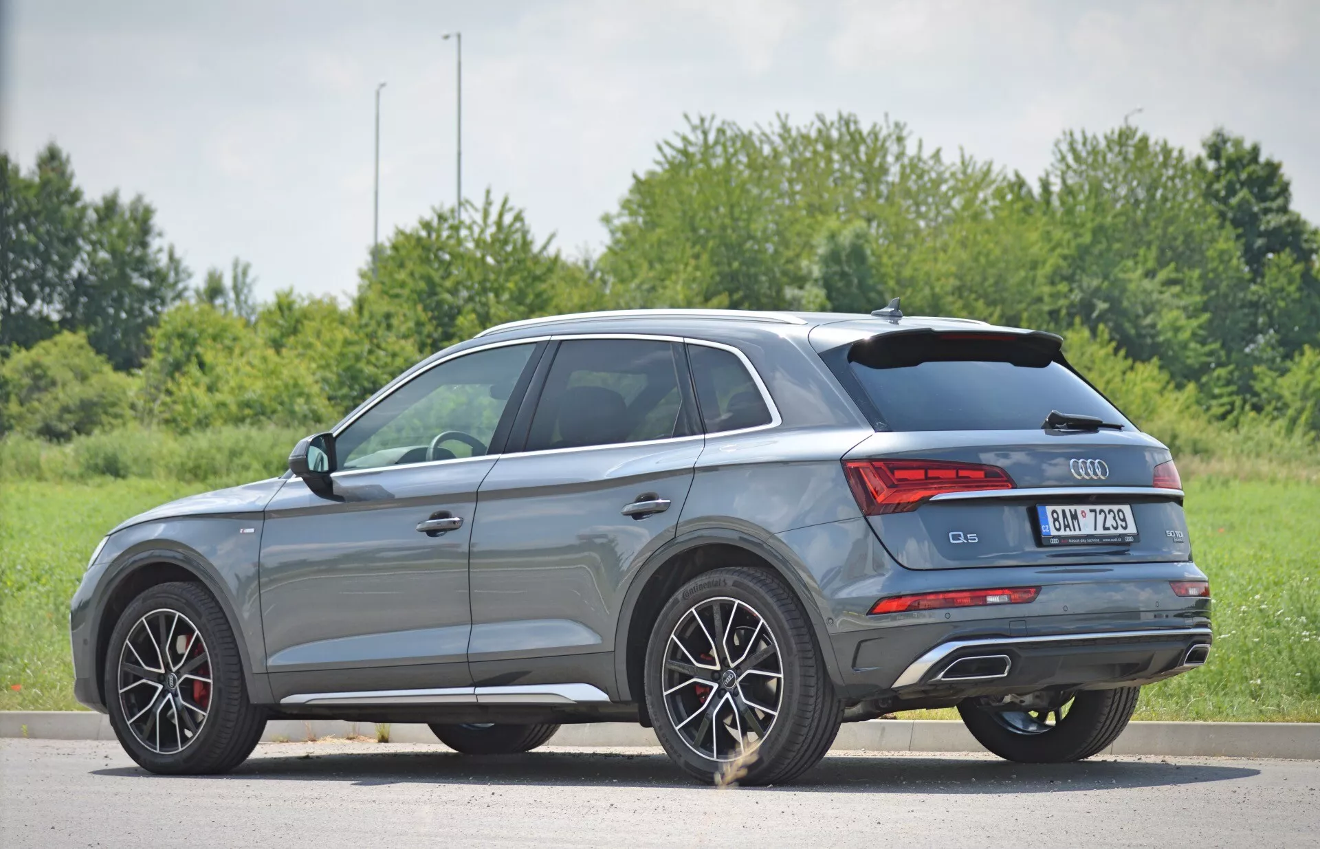 Un SUV Audi Q5 Sportback gris de l'année modèle 2020 est affiché avec une vue arrière trois quarts, mettant en valeur des lignes de carrosserie élégantes, des jantes en alliage multi-branches, des feux arrière à LED, des doubles sorties d'échappement et une ligne de toit arrière inclinée. La voiture est garée sur une surface plane avec un arrière-plan naturel d'herbe et d'arbres, et la lumière naturelle souligne ses éléments de design sportif et son apparence compacte de SUV de taille moyenne.