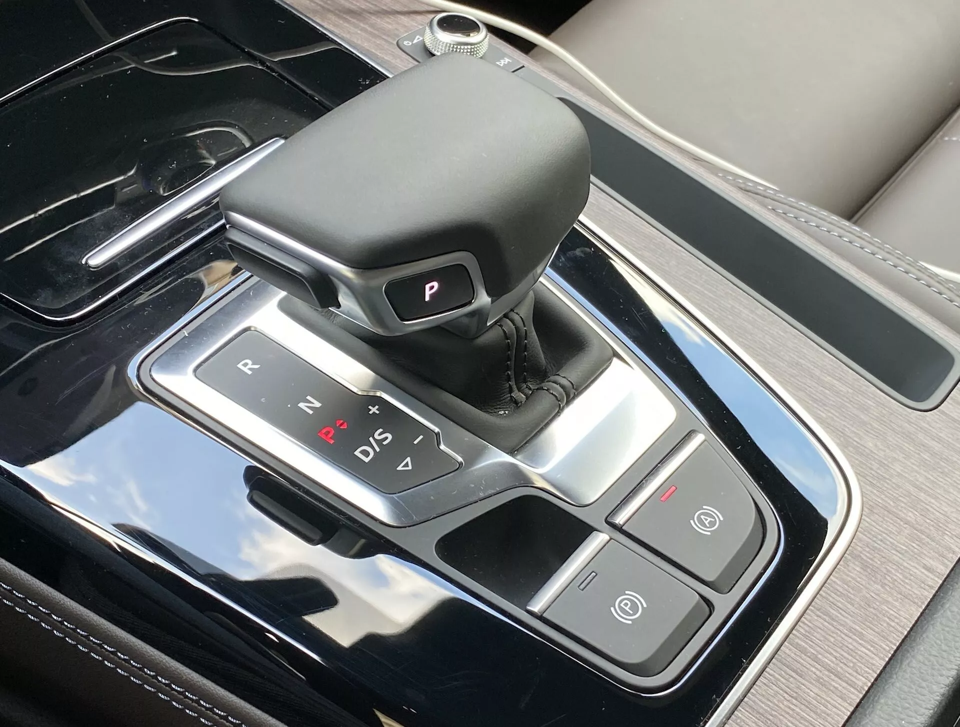 Vue de dessus de la console centrale de l'Audi Q5 Sportback 2020, mettant en évidence la zone raffinée du levier de vitesses avec un sélecteur automatique en cuir, des indicateurs de vitesse illuminés et des boutons de contrôle adjacents pour le frein de stationnement automatique et la fonction Auto Start/Stop. La console présente une finition noire piano avec des garnitures argentées, reflétant le design intérieur luxueux et la technologie moderne.