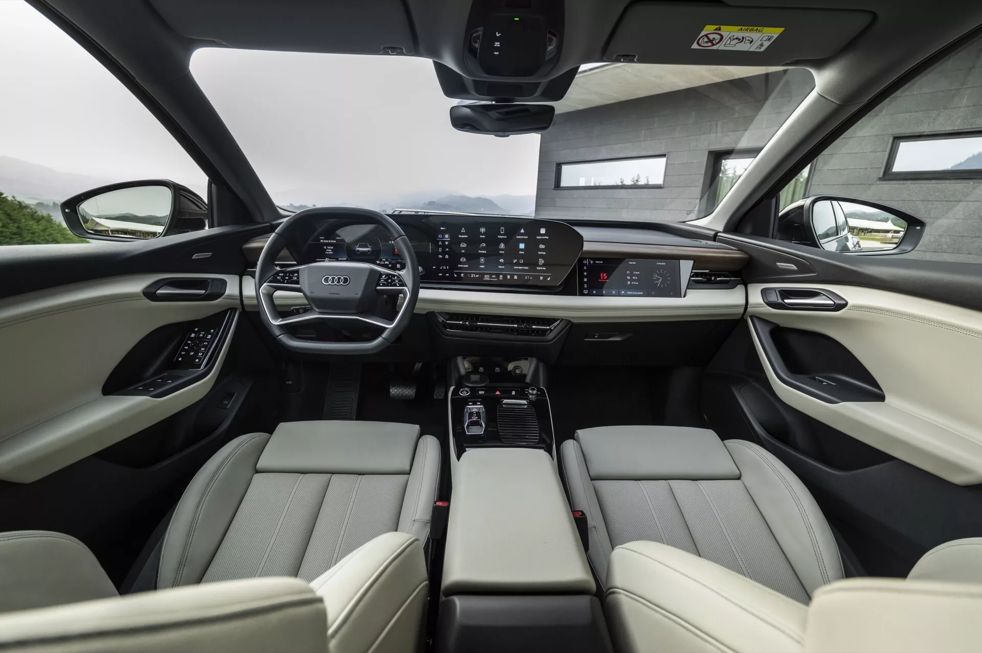 Vue d'intérieur de l'Audi Q6 e-tron 2024 depuis la perspective de la banquette arrière, mettant en avant la rangée avant. Les sièges avant en cuir clair disposent de repose-têtes ajustables et de commandes électroniques. Le tableau de bord présente un large affichage digital et un grand écran tactile central pour l'infodivertissement, entouré de boutons de contrôle pour le climat et les modes de conduite. La console centrale comprend des porte-gobelets, un espace de rangement et un sélecteur de transmission automatique. Le volant est équipé de multiples commandes et le toit panoramique suggère une lumière naturelle abondante. Dans l'ensemble, l'image met en valeur les technologies avancées et les caractéristiques de luxe du véhicule.