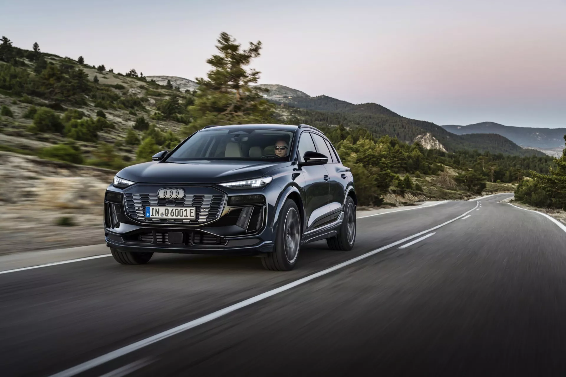 Audi Q6 e-tron 2024 avec un design moderne, une grande calandre avant, des phares LED élégants et des lignes sculptées de la carrosserie. Vue de trois quarts avant avec une prise de vue dynamique en mouvement sur une route vallonnée, mettant en valeur la performance et la maniabilité.