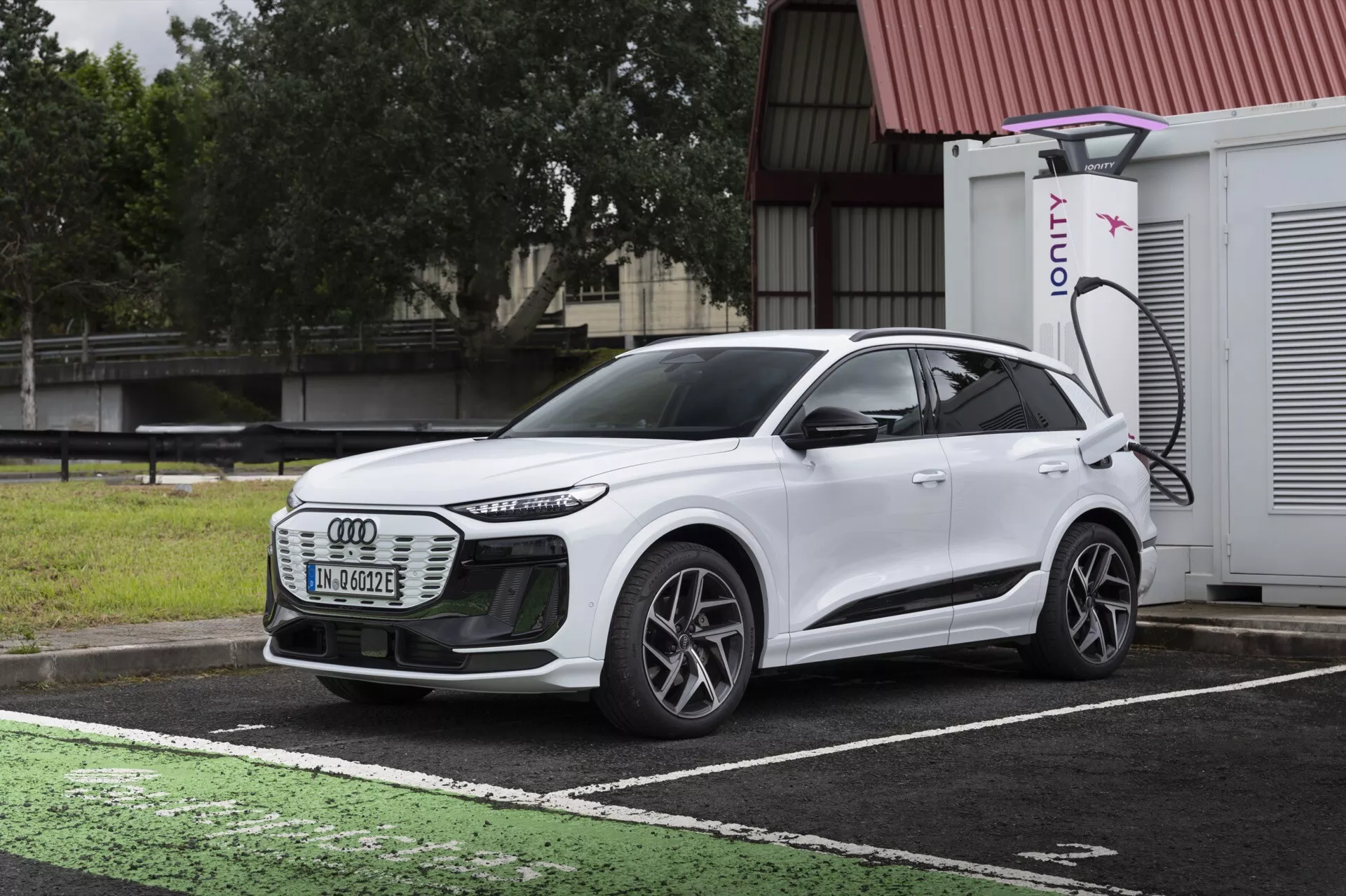 Vue avant trois quarts d'un SUV électrique Audi Q6 e-tron 2024 blanc, mettant en évidence la calandre avant, les phares à LED, les lignes aérodynamiques, les roues à rayons multiples et le profil latéral. Le véhicule est stationné dans un emplacement désigné pour la recharge des véhicules électriques, avec un port de recharge visible et un chargeur IONITY en arrière-plan. L'image montre en contraste le design de la voiture avec un ciel sombre et nuageux et un trottoir mouillé, illustrant la technologie moderne des véhicules électriques et l'éclairage avancé.