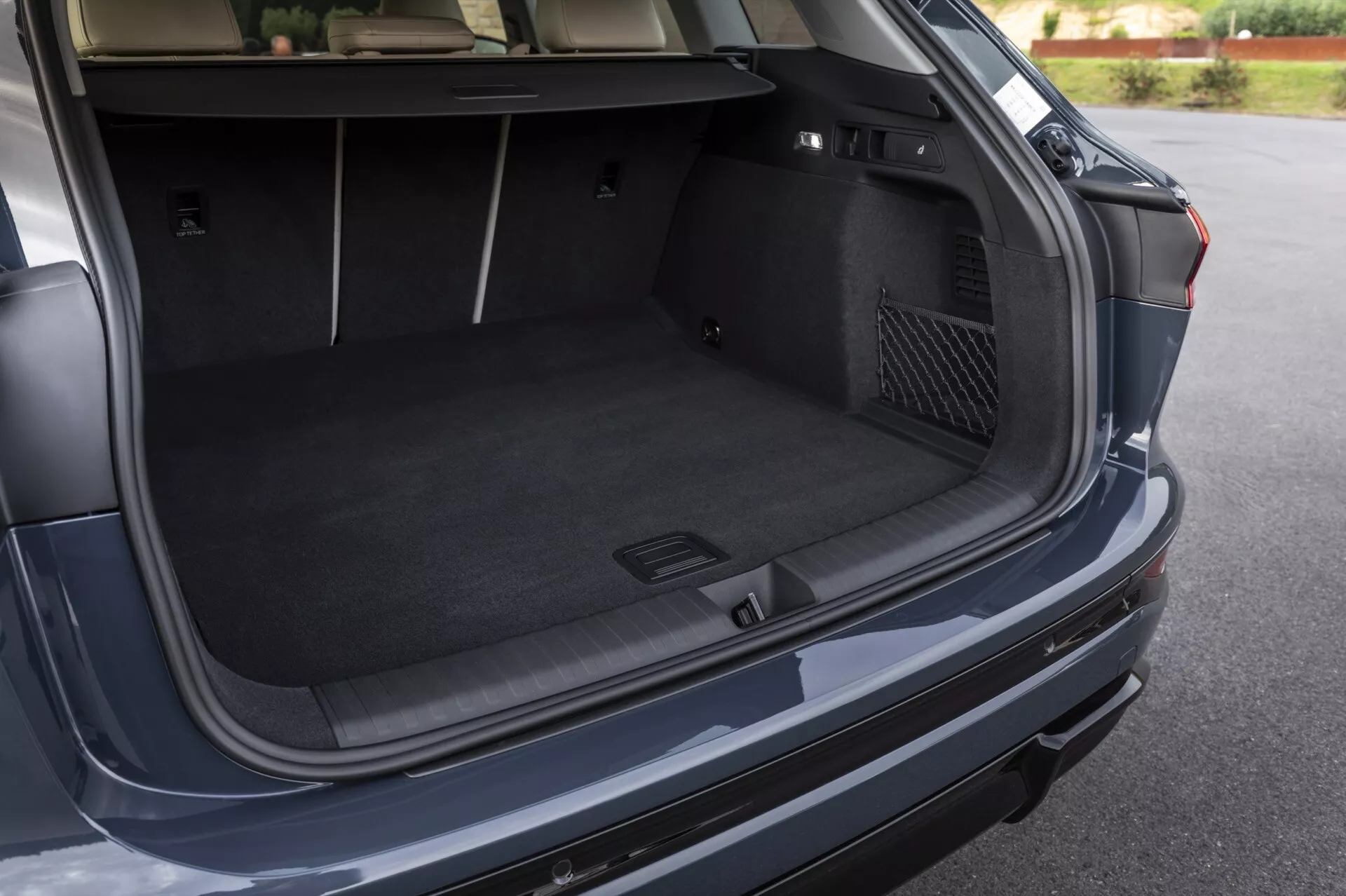 L'image montre le coffre ouvert d'un véhicule moderne Audi Q6 e-tron 2024, capturée sous un angle légèrement au-dessus et à droite de l'arrière de la voiture. Le point de vue offre une vue claire du spacieux espace de chargement recouvert de moquette, mettant en évidence sa profondeur et sa largeur. Les caractéristiques notables incluent un compartiment moulé à gauche pour des petits articles ou de l'équipement d'urgence et une poche de rangement à filet à droite. Un bouton de fermeture automatique possible est visible sur le couvercle du coffre. Le pare-chocs arrière et une vue partielle du feu arrière droit suggèrent la conception extérieure du véhicule.