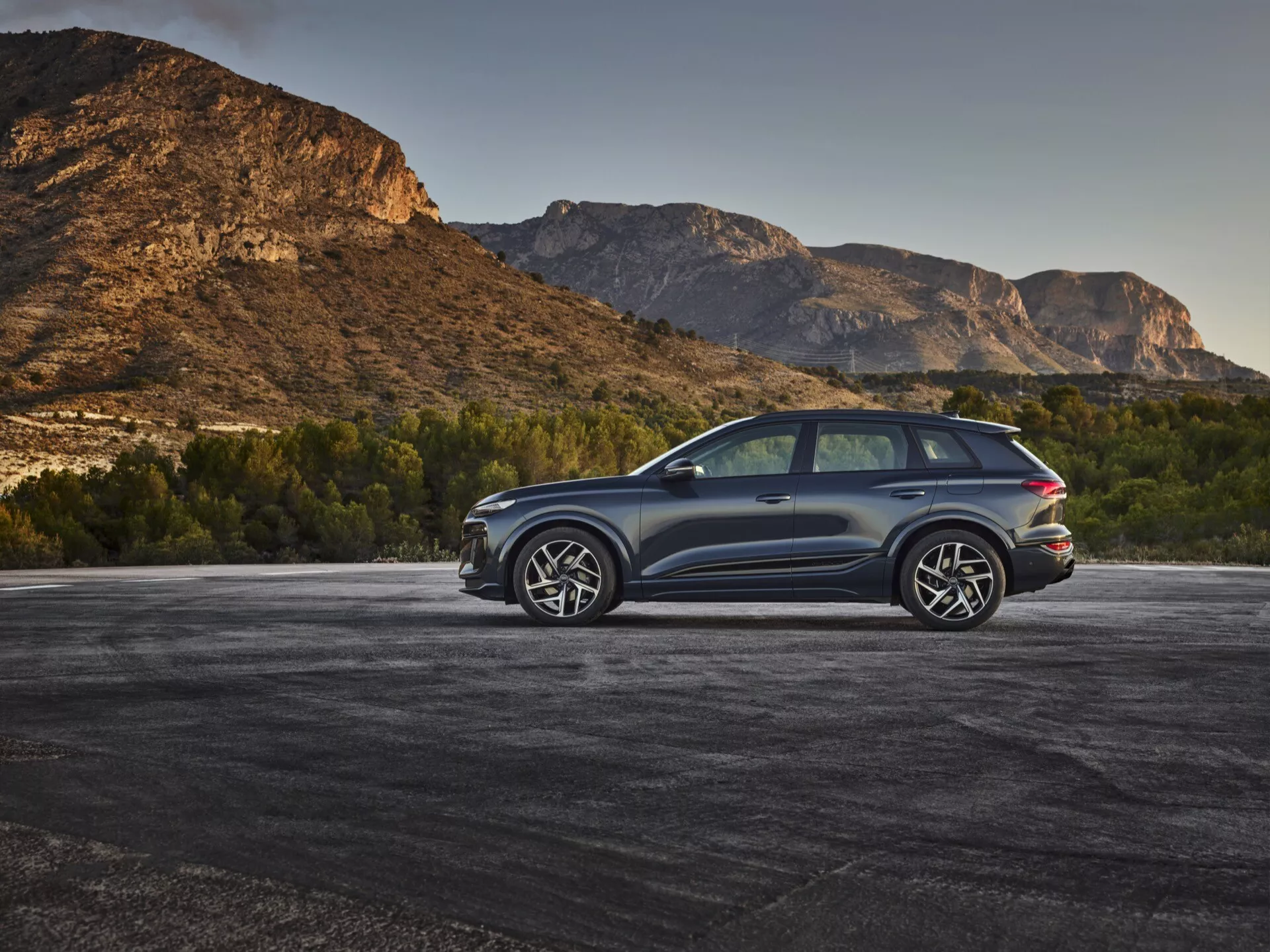 Un SUV moderne, supposé être l'Audi Q6 e-tron 2024, stationné à l'extérieur avec un arrière-plan montagneux, capturé en vue arrière de trois quarts depuis un angle élevé. La couleur métallisée foncée de la carrosserie contraste avec l'environnement naturel, mettant en valeur des lignes élégantes et des contours aérodynamiques. Les détails visibles incluent des feux arrière à LED, de grandes roues en alliage à plusieurs rayons, un système d'échappement double, des vitres teintées, un toit panoramique et des moulures de protection le long des bords inférieurs. La lunette arrière est légèrement inclinée avec un spoiler arrière et des rétroviseurs latéraux de grande taille, probablement dotés de clignotants intégrés, renforçant l'attrait sportif et luxueux du véhicule.