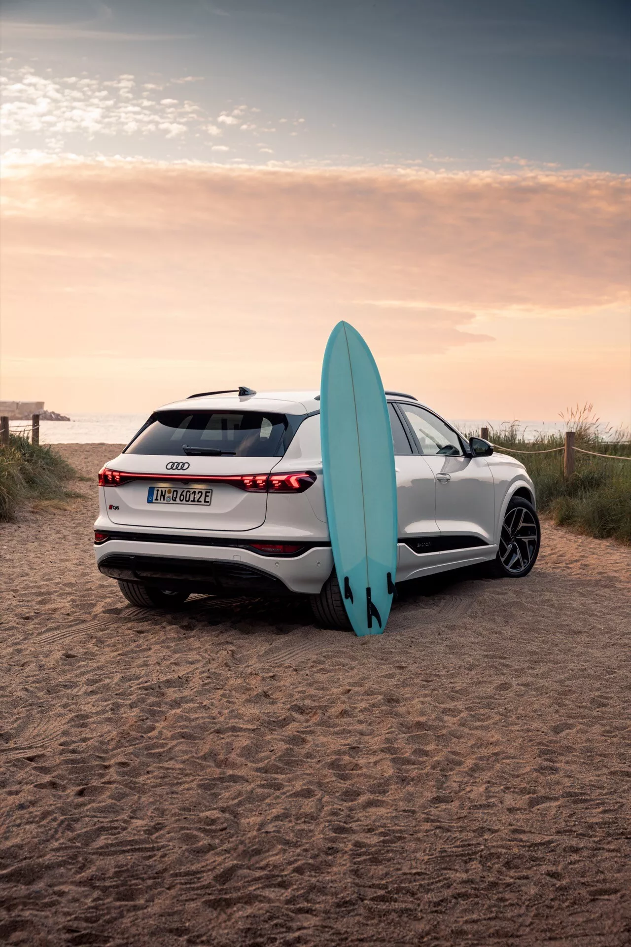 L'image montre un SUV Audi Q6 e-tron 2024 blanc garé sur un terrain sablonneux de plage avec une planche de surf. Photographié sous un angle de trois quarts arrière, il capture les lignes élégantes de la voiture, les feux arrière à LED, les capteurs de stationnement, la caméra de recul et les roues en alliage multi-branches. Le reflet du coucher de soleil met en valeur la qualité avancée de la peinture, tandis que la planche de surf appuyée contre la voiture souligne un élément de style de vie aventureux.