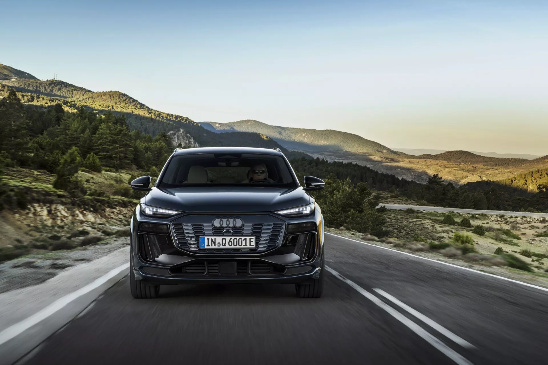 Vue frontale de l'Audi Q6 e-tron 2024, montrant sa calandre, ses phares et son pare-chocs avec le logo des quatre anneaux entrelacés. La plaque d'immatriculation de style européen est montée sur la calandre et la désignation 'e-tron' indique sa nature électrique/hybride. La voiture roule sur une route ouverte avec un paysage montagneux en arrière-plan, suggérant une configuration de fin d'après-midi. Les caractéristiques incluent des phares LED élégants, une calandre esthétique, des capteurs/caméras d'assistance au conducteur, des rétroviseurs aérodynamiques et un design efficace.