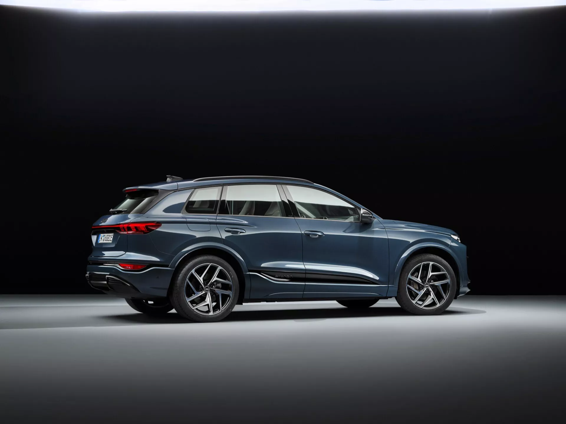 Prise de vue de profil en contre-plongée de l'Audi Q6 e-tron 2024 dans un studio avec fond sombre, avec des roues en alliage à plusieurs rayons sportives, un design de carrosserie fluide avec une ligne de caractère, des rétroviseurs aérodynamiques avec clignotants intégrés, des encadrements de fenêtres chromés, un toit panoramique, des rails de toit, une hauteur de conduite modérée et un système d'échappement double intégré.