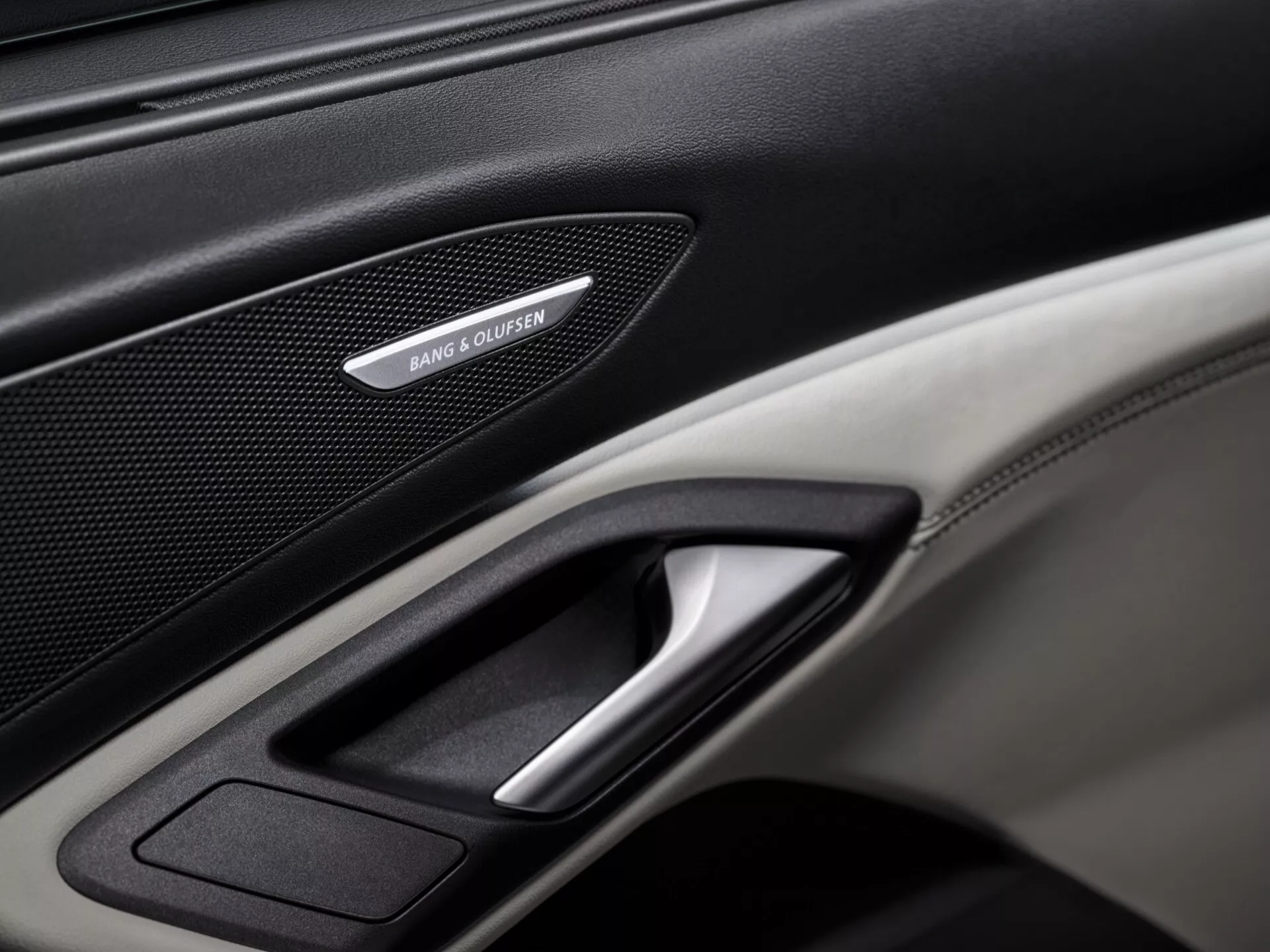 Photographie en gros plan du panneau de porte intérieur de l'Audi Q6 e-tron 2024 GF, mettant en évidence des caractéristiques haut de gamme telles que la grille de haut-parleur Bang & Olufsen, la poignée de porte chromée et une combinaison de textures en cuir et en fibre de carbone. L'image bien éclairée se concentre sur les détails complexes du panneau, mettant en valeur les éléments de luxe et de technologie du véhicule.