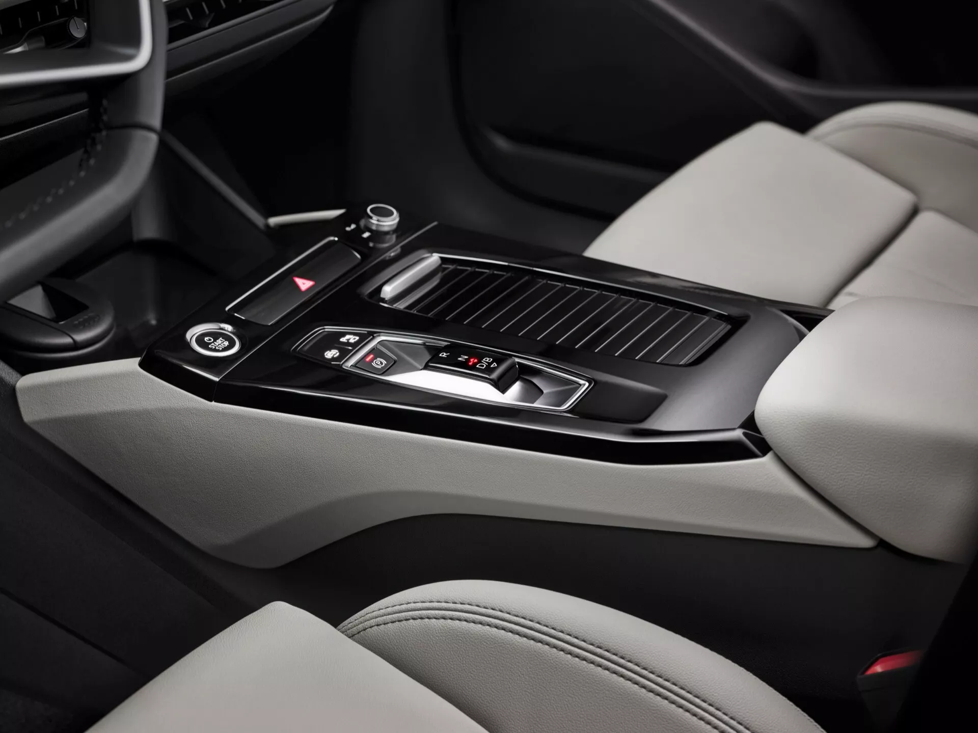 Photo de l’intérieur de l’Audi Q6 e-Tron 2024, axée sur la console centrale. L’image, prise de la perspective du passager, met en évidence un levier de vitesses noir brillant avec des détails métalliques, plusieurs boutons de commande, un petit espace de rangement avec un couvercle coulissant, deux porte-gobelets, un accoudoir en cuir, des bouches d’aération pour le contrôle climatique, et des parties de sièges en cuir clair avec des sections contrastantes plus sombres. Les caractéristiques de la console centrale montrent un design moderne et des éléments luxueux.