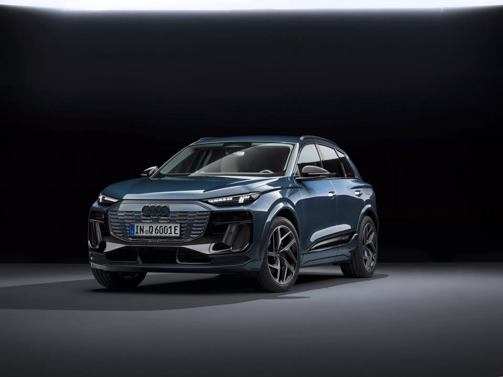 L'image montre un SUV Audi Q6-e-tron 2024 moderne vu de trois quarts avant, mettant en évidence à la fois son profil avant et latéral. Les principaux identificateurs incluent le badge Audi à quatre anneaux sur la calandre et le badge 'e-tron' indiquant sa nature électrique. La prise de vue est effectuée d'une perspective basse, accentuant la stature du véhicule et montrant ses lignes de conception, les passages de roues musclés et les caractéristiques aérodynamiques. La voiture est centrée dans le cadre, placée sur une surface réfléchissante lisse avec un éclairage doux et diffus et un fond de dégradé sombre, mettant en valeur le design du véhicule. Les caractéristiques technologiques visibles incluent des phares à LED, des feux diurnes (DRL), un écran numérique et des roues en alliage au design complexe. La présentation générale soignée suggère une photographie promotionnelle mettant en avant la technologie, la conception et les aspects luxueux du véhicule.