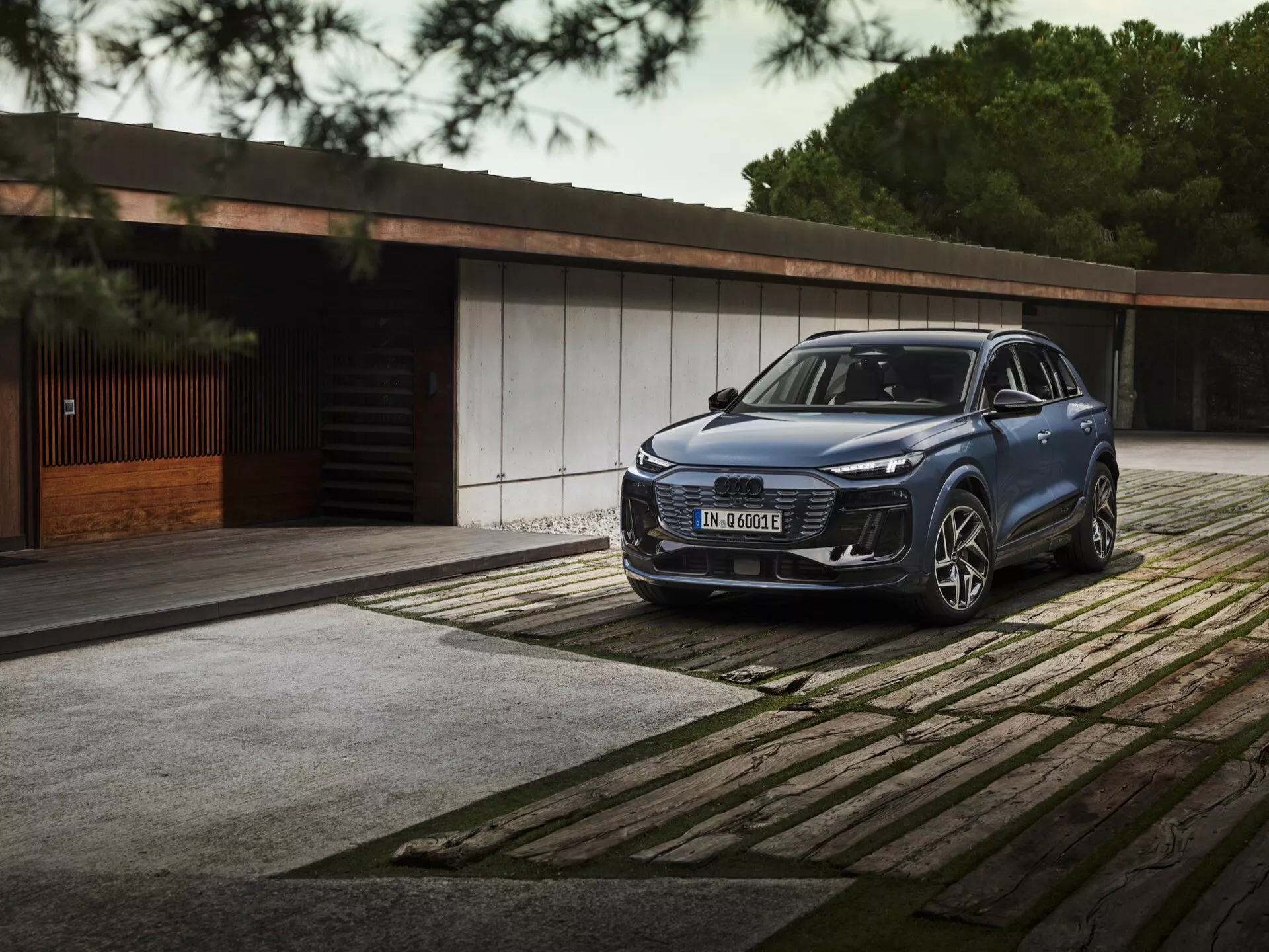 Vue trois quarts avant du SUV Audi Q6 E-Tron 2024 avec des phares LED élégants, une calandre proéminente et un grand emblème garé sur un pavé à motifs. Bâtiment de luxe et arbres en arrière-plan soulignent le luxe et l'exclusivité.