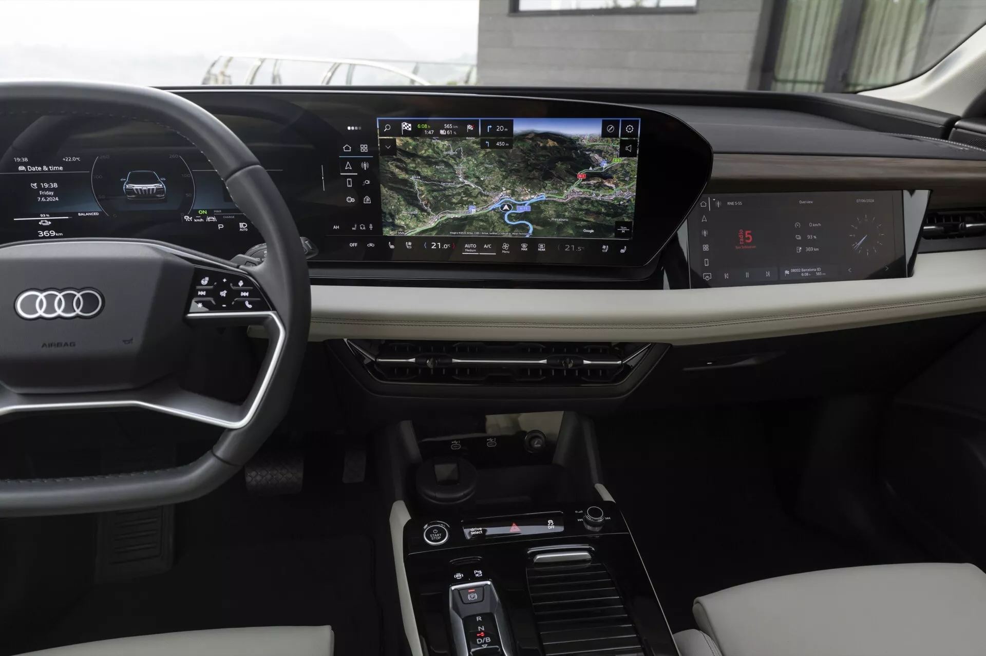 L'intérieur d'une moderne Audi Q6 e-tron 2024, vu du point de vue du conducteur. Les caractéristiques incluent un volant à trois branches en cuir avec des commandes intégrées, un tableau de bord numérique affichant la vitesse et la navigation, et un système d'infodivertissement à écran tactile horizontal. La console centrale comprend des commandes pour les réglages climatiques et les modes de conduite. Des matériaux luxueux comme le cuir et le bois rehaussent l'esthétique.