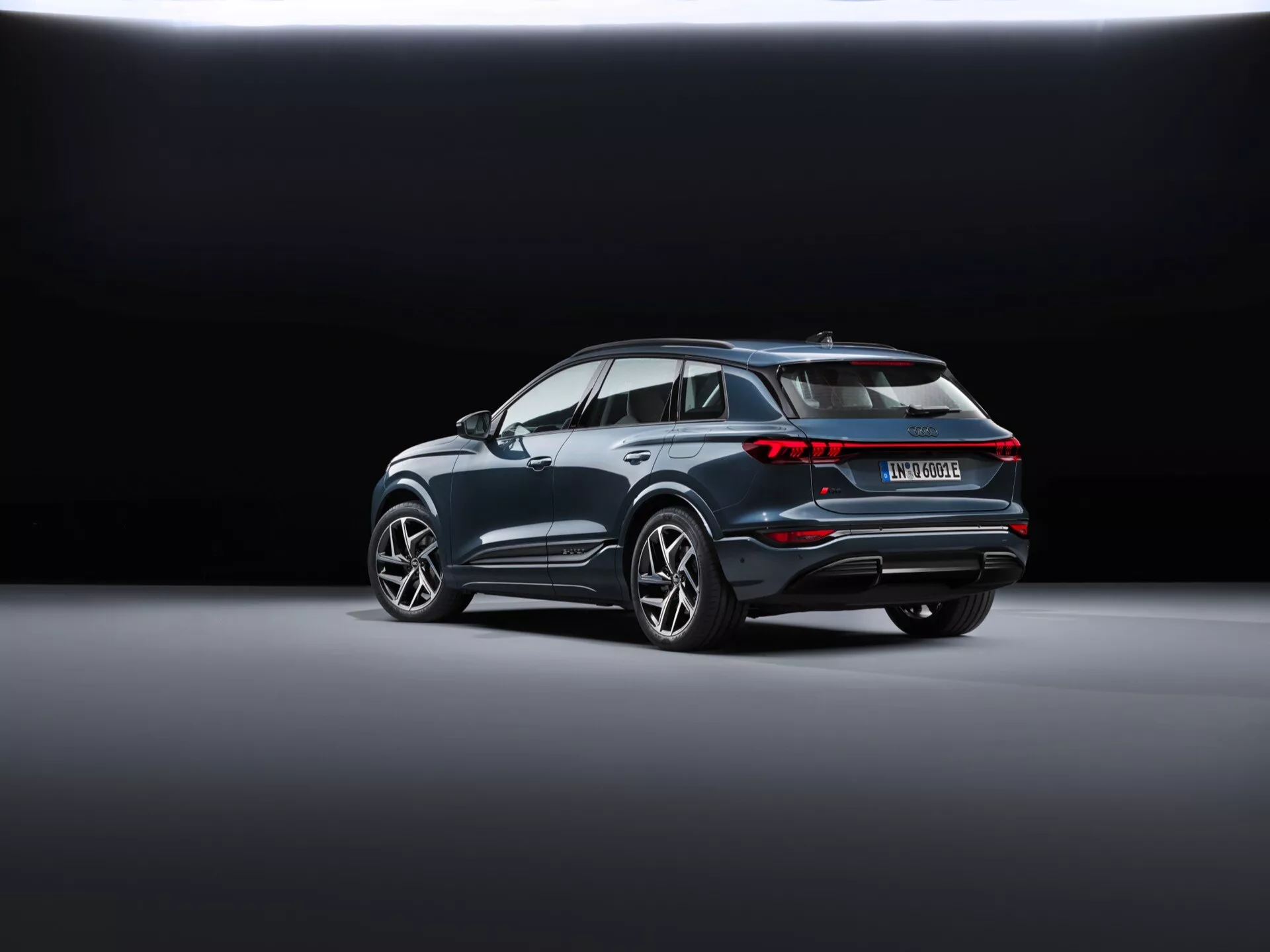 La vue arrière trois-quarts de l'Audi Q6 e-tron 2024 (gf) dévoile un SUV moderne positionné en diagonale sur un sol de showroom lisse et réfléchissant. L'arrière-plan passe du foncé au clair, mettant en valeur le véhicule. Les caractéristiques visibles incluent un assemblage de feux arrière interconnectés, un aileron de toit distinctif, des arches de roue prononcées avec de grandes jantes en alliage, une garniture chromée le long des fenêtres, un pare-chocs arrière avec des réflecteurs et des capteurs et un système d'échappement double suggérant des performances, ce qui indique qu'il pourrait s'agir d'un modèle hybride ou électrique.