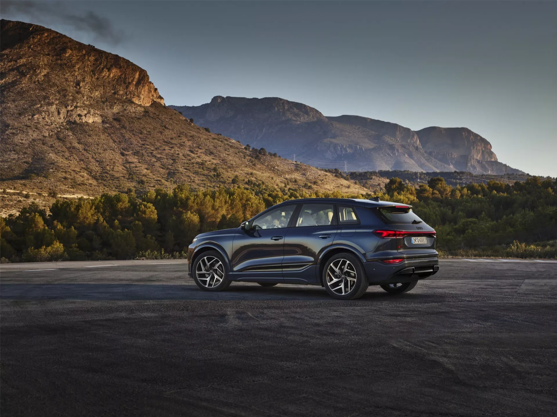 L'image montre un SUV moderne Audi Q6 e-Tron 2024 dans un cadre extérieur avec un arrière-plan pittoresque de montagnes au lever ou au coucher du soleil. L'angle arrière de trois quarts met en évidence les feux arrière à LED distinctifs, le design propre du hayon pouvant intégrer une caméra de recul et un système d'échappement double. Le profil latéral présente des jantes en alliage à plusieurs branches, de grandes fenêtres et une ligne de toit inclinée, suggérant une finition sportive ou de luxe avec d'éventuelles caractéristiques aérodynamiques. Les rétroviseurs latéraux pourraient intégrer une technologie de surveillance des angles morts. L'éclairage naturel renforce l'attrait du véhicule, l'associant à l'aventure et à la capacité.