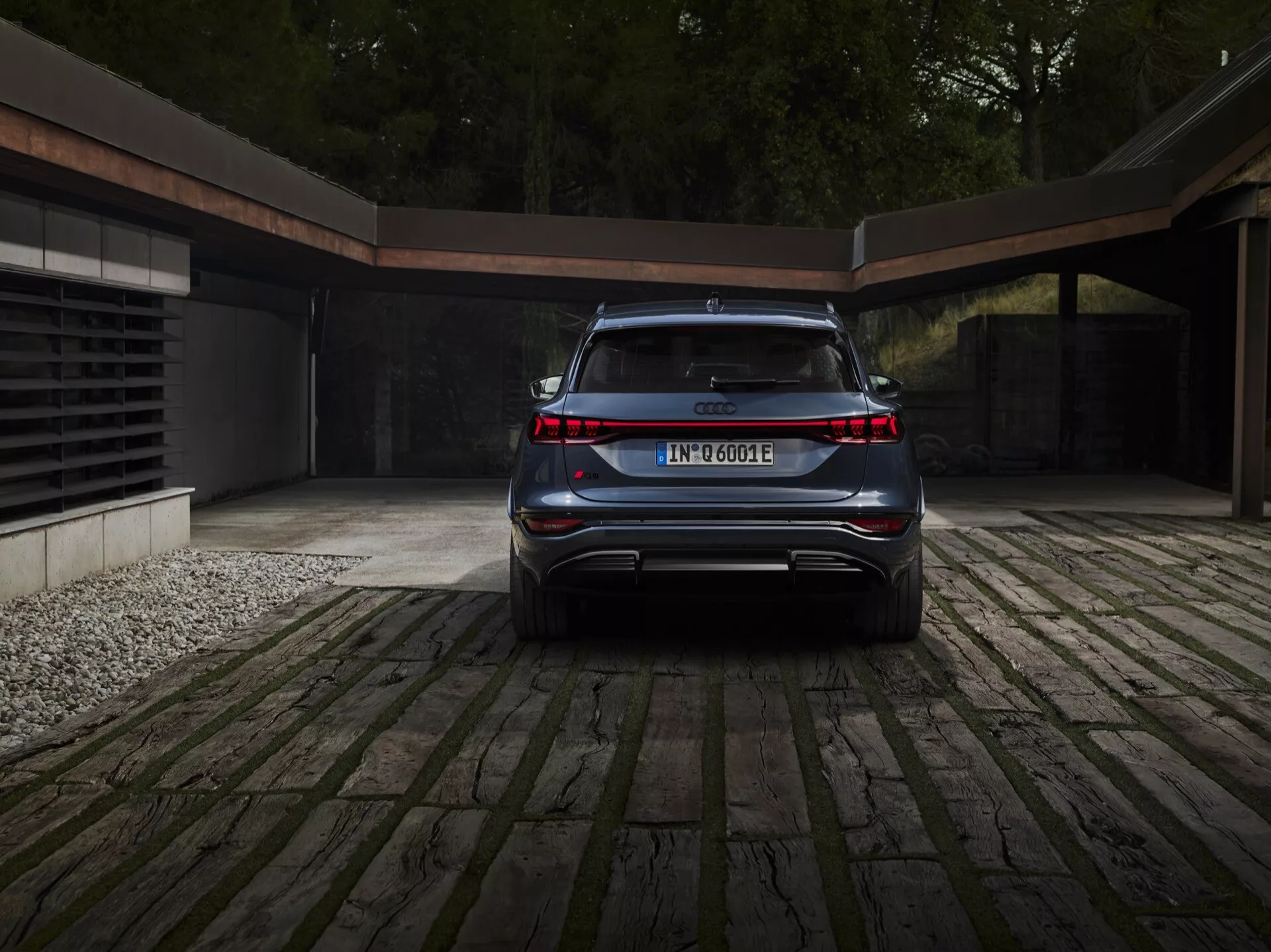 Vue de l'arrière de l'Audi Q6 e-tron 2024 mettant en valeur son design propre, élégant et moderne. Les caractéristiques clés visibles sont la bande lumineuse continue reliant les deux feux arrière, le badge du modèle et du groupe motopropulseur électrique, le pare-chocs arrière intégré avec diffuseur sportif, la lunette arrière inclinée vers l'avant suggérant un SUV ou crossover, les sorties d'échappement cachées et la plaque d'immatriculation centrée. La voiture est garée sur une allée pavée avec un aperçu d'un garage contemporain en arrière-plan, indiquant un style de vie luxueux et respectueux de l'environnement.
