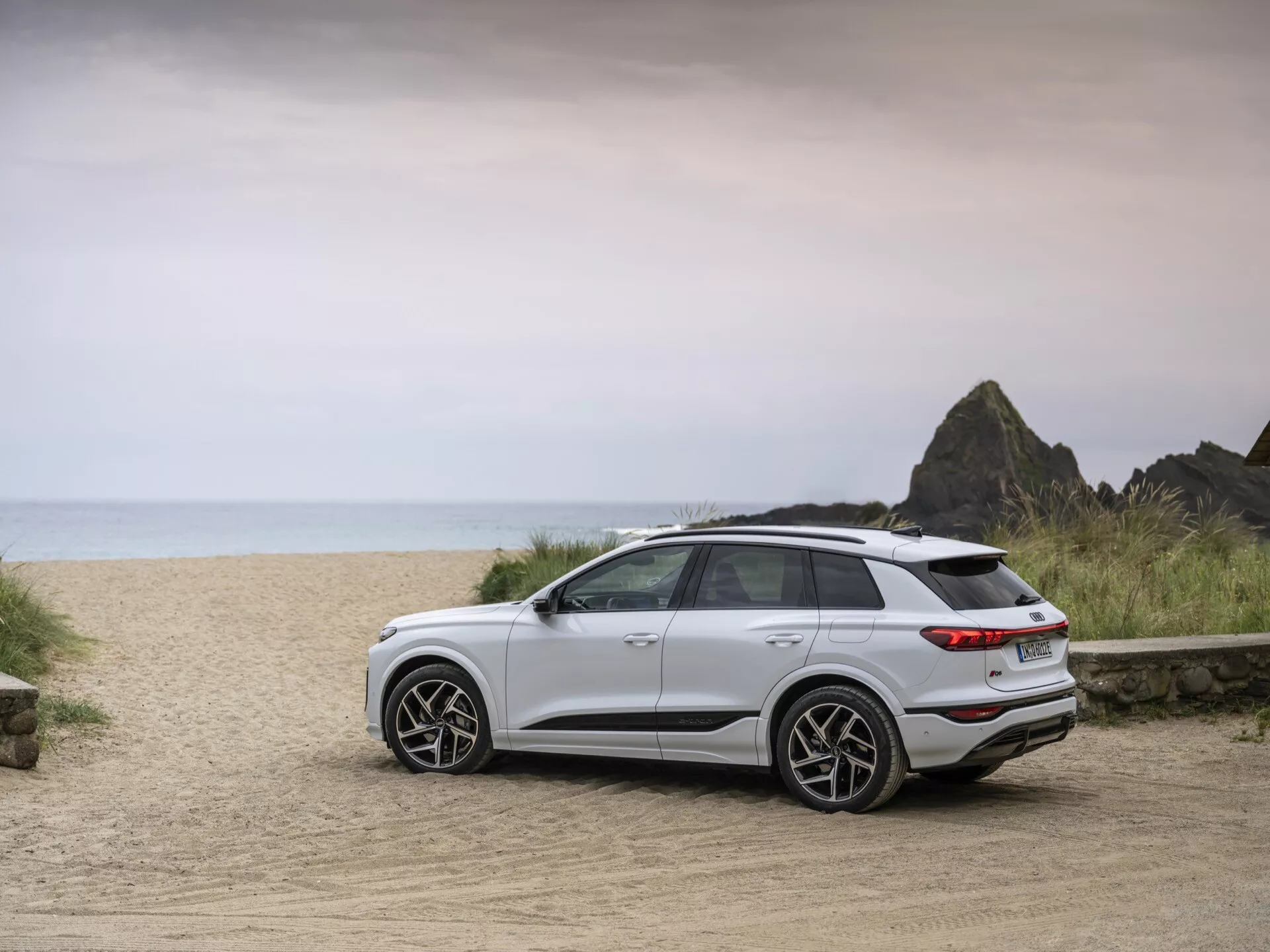Un SUV blanc Audi Q6 e-tron 2024 est garé sur une route de terre avec un fond de plage, capturé sous un angle trois-quarts arrière. L'image met en valeur les éléments de design modernes du véhicule, y compris les feux arrière, le pare-chocs arrière, les roues élégantes et les lignes épurées. La garde au sol élevée et la posture générale de la voiture indiquent son aptitude à divers terrains. L'éclairage doux et homogène suggère un jour nuageux ou proche du lever/coucher du soleil, renforçant le cadre aventureux. Des roues en alliage modernes, un spoiler arrière et des feux arrière LED sont visibles, signalant une technologie avancée et un caractère sportif.