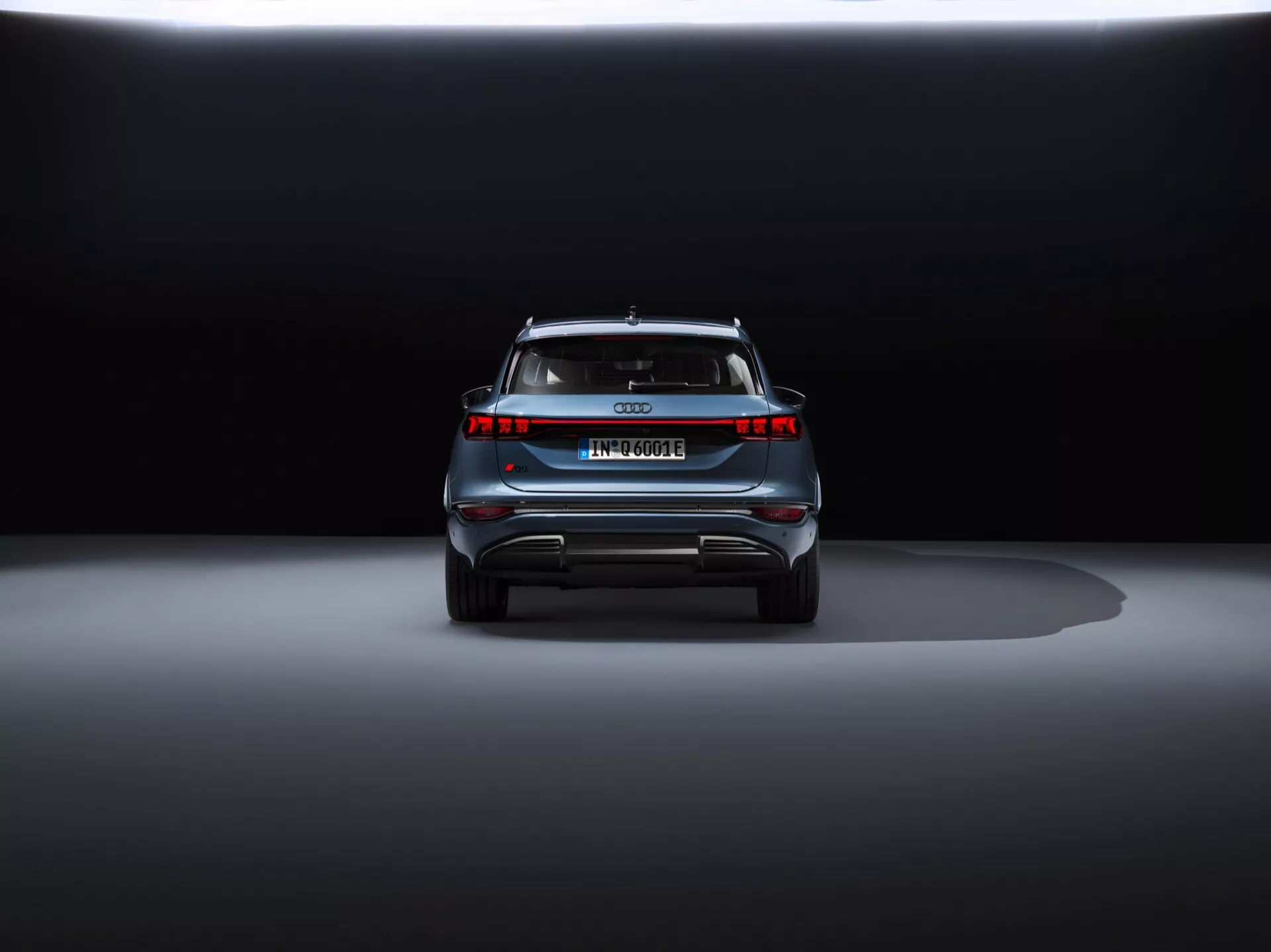 Vue arrière de l'Audi Q6 e-tron 2024 en exposition avec éclairage dramatique. Prise de vue au niveau du sol mettant en évidence les feux arrière à LED modernes, l'absence de tuyaux d'échappement, une barre de lumière pleine largeur, une plaque d'immatriculation de style européen, un spoiler de toit et des éléments de sécurité réfléchissants, indiquant qu'il s'agit d'un véhicule de luxe électrique ou hybride