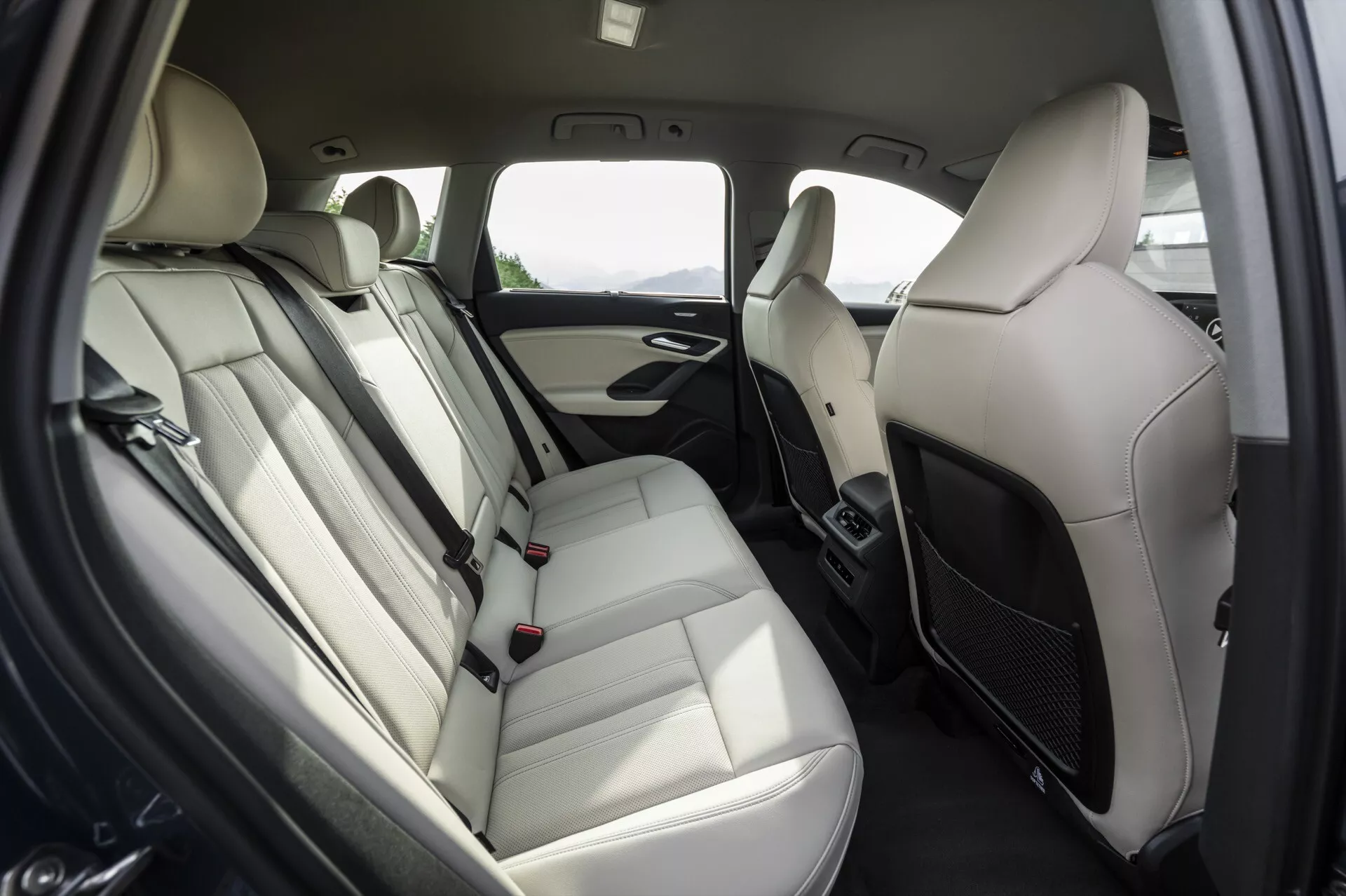 Compartiment arrière de la 2024 Audi Q6 e-tron axé sur l'agencement des sièges arrière, banquettes en cuir clair avec appuie-têtes, accoudoir et ceintures de sécurité visibles avec boucles. Panneau de porte avec grille de haut-parleur, poignée et éventuels commandes de fenêtre. Schéma de couleurs bicolores avec des garnitures claires et foncées. Bouches d'aération entre les sièges avant indiquant un contrôle climatique arrière. Design moderne, luxueux et confortable pour les passagers.