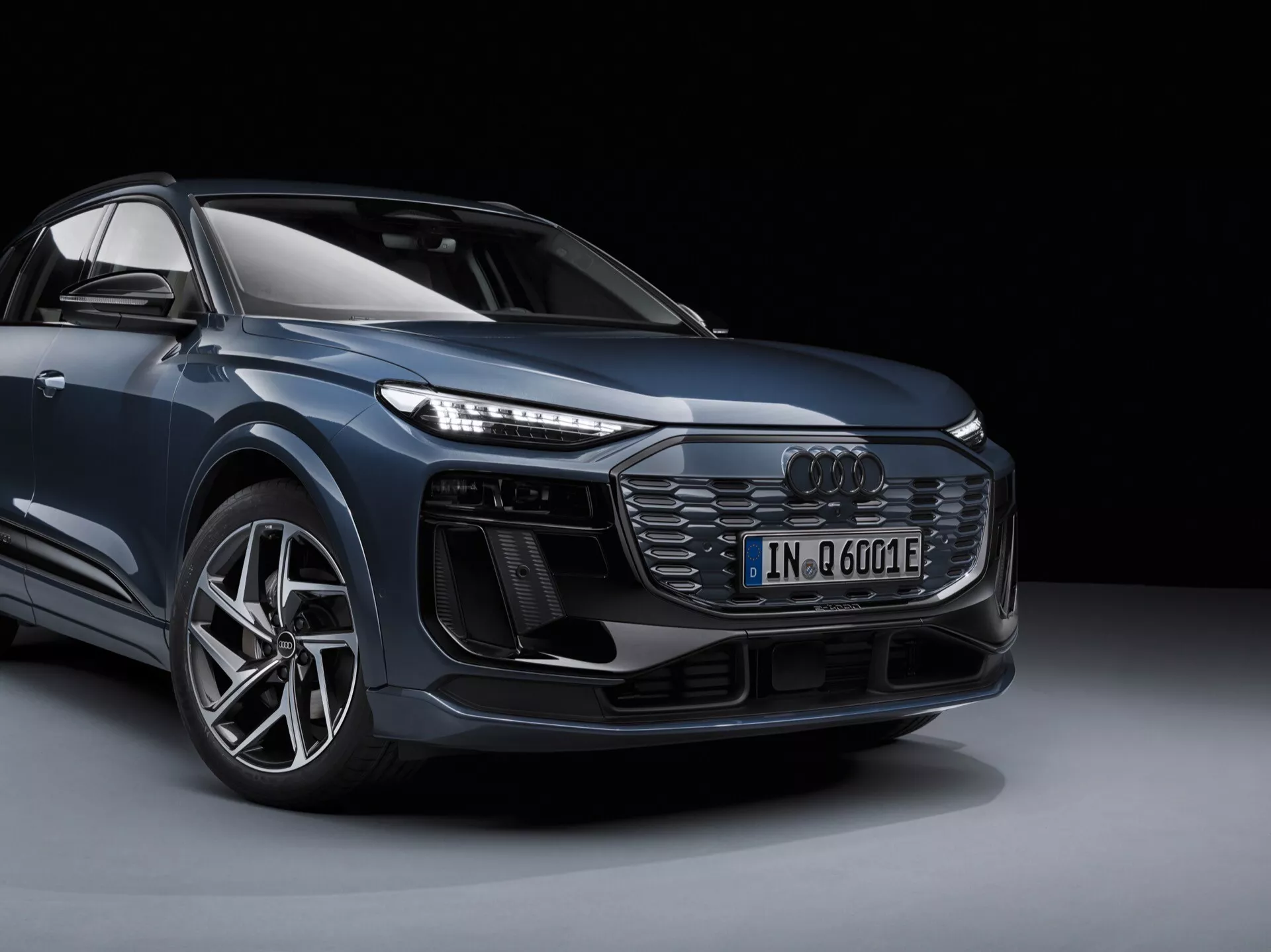 L'image présente une Audi Q6 e-tron 2024 moderne dans un cadre de studio, vue depuis un angle de trois quarts avant fournissant une impression claire du design du véhicule de face et de côté. Le coin avant gauche de la voiture est clairement visible, incluant la grille avant avec un motif en nid d'abeilles et l'emblème Audi situé au centre, des phares LED complexes, des prises d'air supplémentaires ou des capteurs sous les phares, un pare-chocs avant sportif, une grande roue en alliage avec un pneu à profil bas et le contour d'un rétroviseur latéral. L'image met en avant le badge du modèle électrifié ('e') et souligne ses caractéristiques de design et technologiques. L'arrière-plan est simple et sombre, sans distractions par rapport à la voiture elle-même.