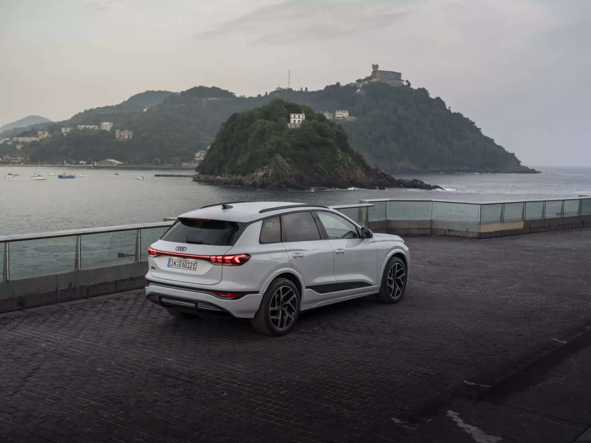 Vue arrière trois-quarts d'un SUV Audi Q6 e-tron 2024 blanc moderne garé sur une esplanade en bord de mer au crépuscule, montrant ses feux arrière horizontaux à LED, l'écusson du fabricant, l'essuie-glace arrière, les roues en alliage à cinq branches, les rétroviseurs avec clignotants intégrés, les barres de toit et les lignes sculptées de la carrosserie contre un décor serein avec une petite île et un bâtiment, soulignant un mélange d'utilité et de performance.