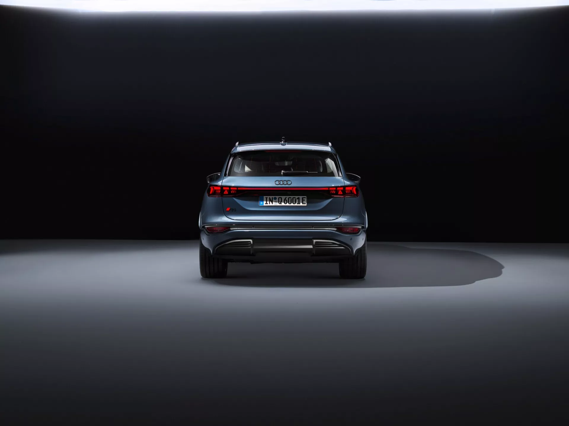 Vue arrière de l'Audi Q6 e-tron 2024 sur un fond sombre, mettant en valeur son design élégant, l'emblème central, le nom du modèle, la barre lumineuse horizontale, le spoiler arrière et le pare-chocs de type diffuseur avec des éléments réfléchissants.