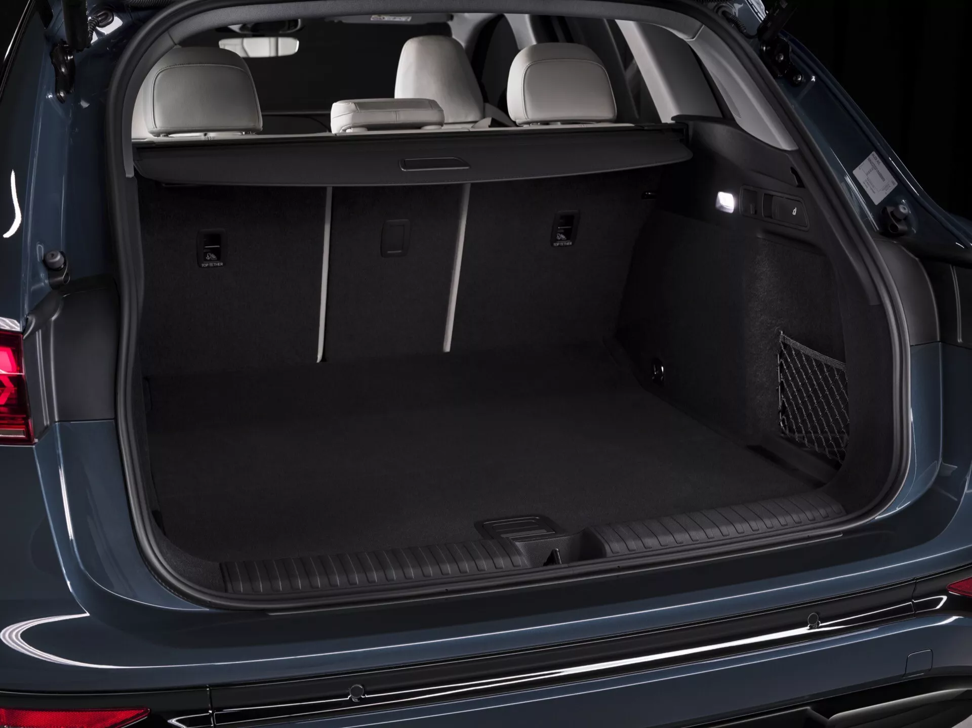 Gros plan du coffre ouvert de l'Audi Q6 e-tron 2024 en perspective arrière, montrant son spacieux espace de chargement. Les fonctionnalités incluent des points d'ancrage, une prise 12 volts, un cache-bagages rétractable, des compartiments de rangement latéraux, un éclairage intérieur et une poche de rangement en filet. L'intérieur est fini avec un tapis sombre et les sièges arrière sont en position verticale, soulignant la capacité et la polyvalence du coffre.