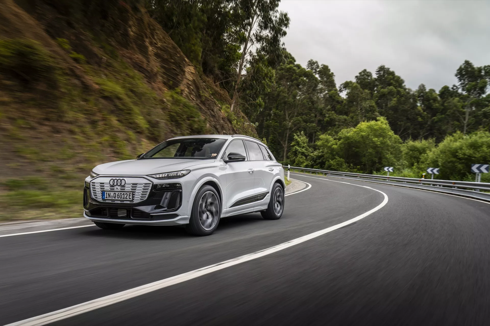 L'image capture une vue latérale dynamique d'un SUV blanc moderne, probablement un modèle Audi Q6 e-tron 2024, roulant sur une route bordée par une zone boisée. L'accent est mis sur la calandre avant, les phares, le profil latéral, les roues et l'aérodynamisme des rétroviseurs latéraux du véhicule. Les phares sont élégants avec une technologie LED, suggérant un éclairage avancé. Les roues en alliage avec un design à plusieurs rayons suggèrent des capacités hautes performances. Le mouvement de la voiture sur une route courbe met en évidence ses capacités de maniabilité et les éventuels systèmes avancés d'assistance à la conduite. L'arrière-plan flou souligne la vitesse et l'efficacité dans la navigation dans des situations difficiles.