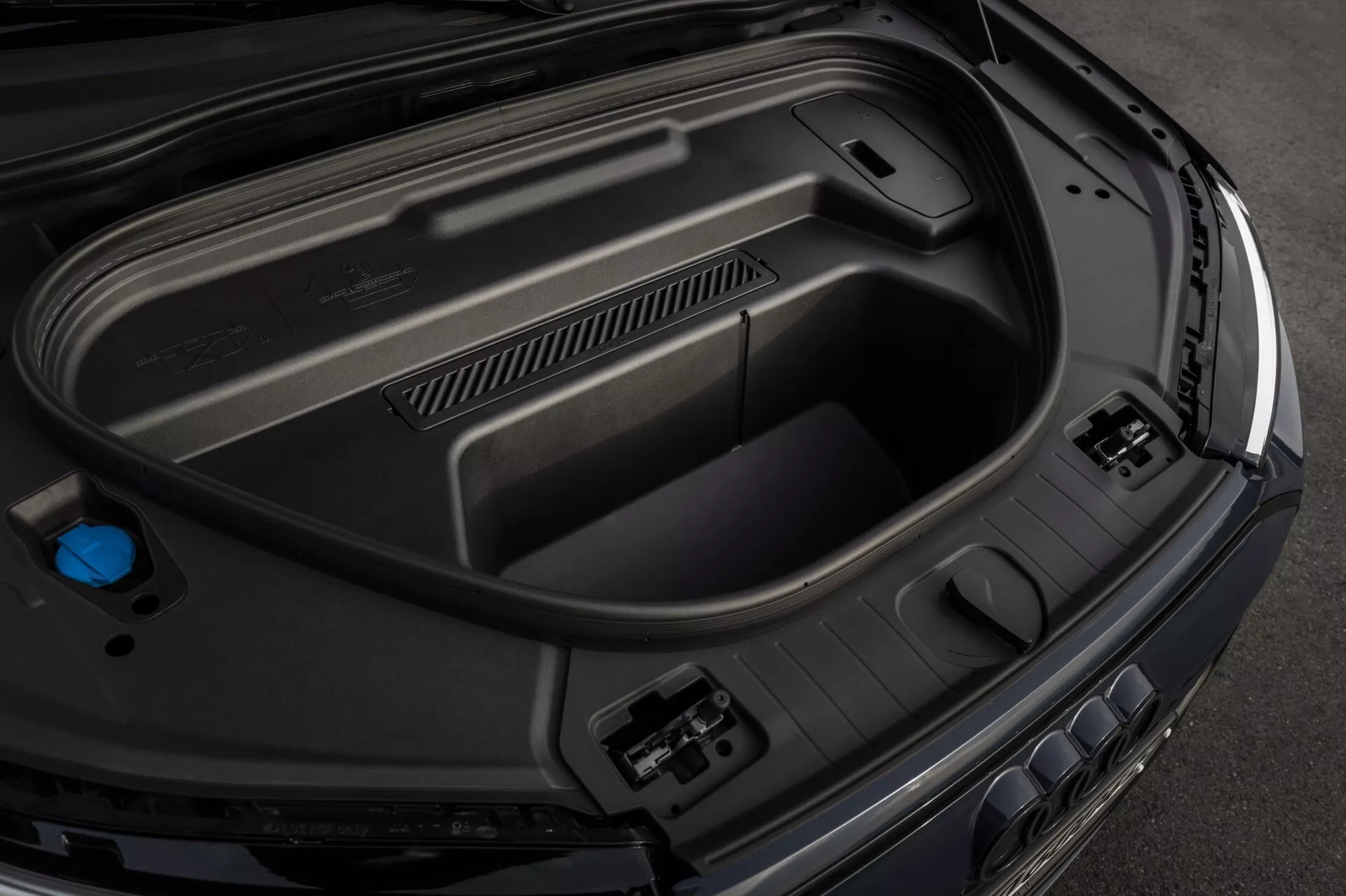 Vue en gros plan sous un angle élevé du compartiment arrière du coffre de l'Audi Q6-e-Tron 2024, mettant en évidence les caractéristiques intérieures, y compris les panneaux en plastique moulé pour la durabilité, les prises d'accessoires de puissance sur le côté droit, le compartiment de rangement encastré au bas et un capuchon bleu à gauche, témoignant d'une technologie automobile avancée et d'une finition haut de gamme et élégante.