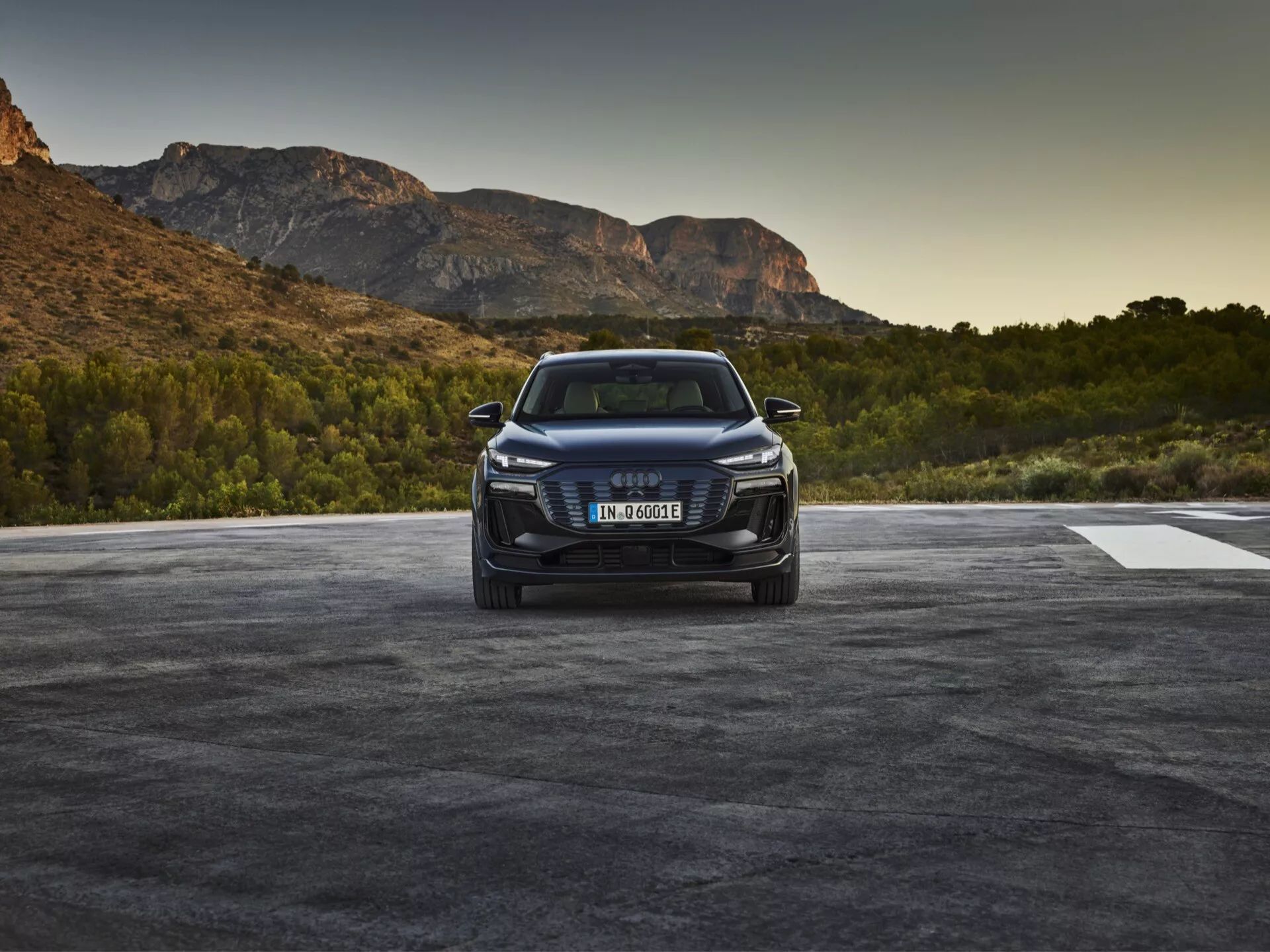 Image d'une Audi Q6 e-tron 2024 photographiée sous un angle de trois quarts avant, montrant la calandre du véhicule avec l'emblème de la marque, des phares à LED ou à laser aiguisés et un pare-chocs avant détaillé avec des prises d'air et possiblement des capteurs ou des caméras indiquant des capacités d'assistance à la conduite. Le capot présente des lignes de contour, et le profil latéral élégant suggère l'aérodynamisme, avec une posture élevée de SUV. La voiture est placée dans un cadre extérieur avec un paysage naturel évoquant le lever ou le coucher du soleil.