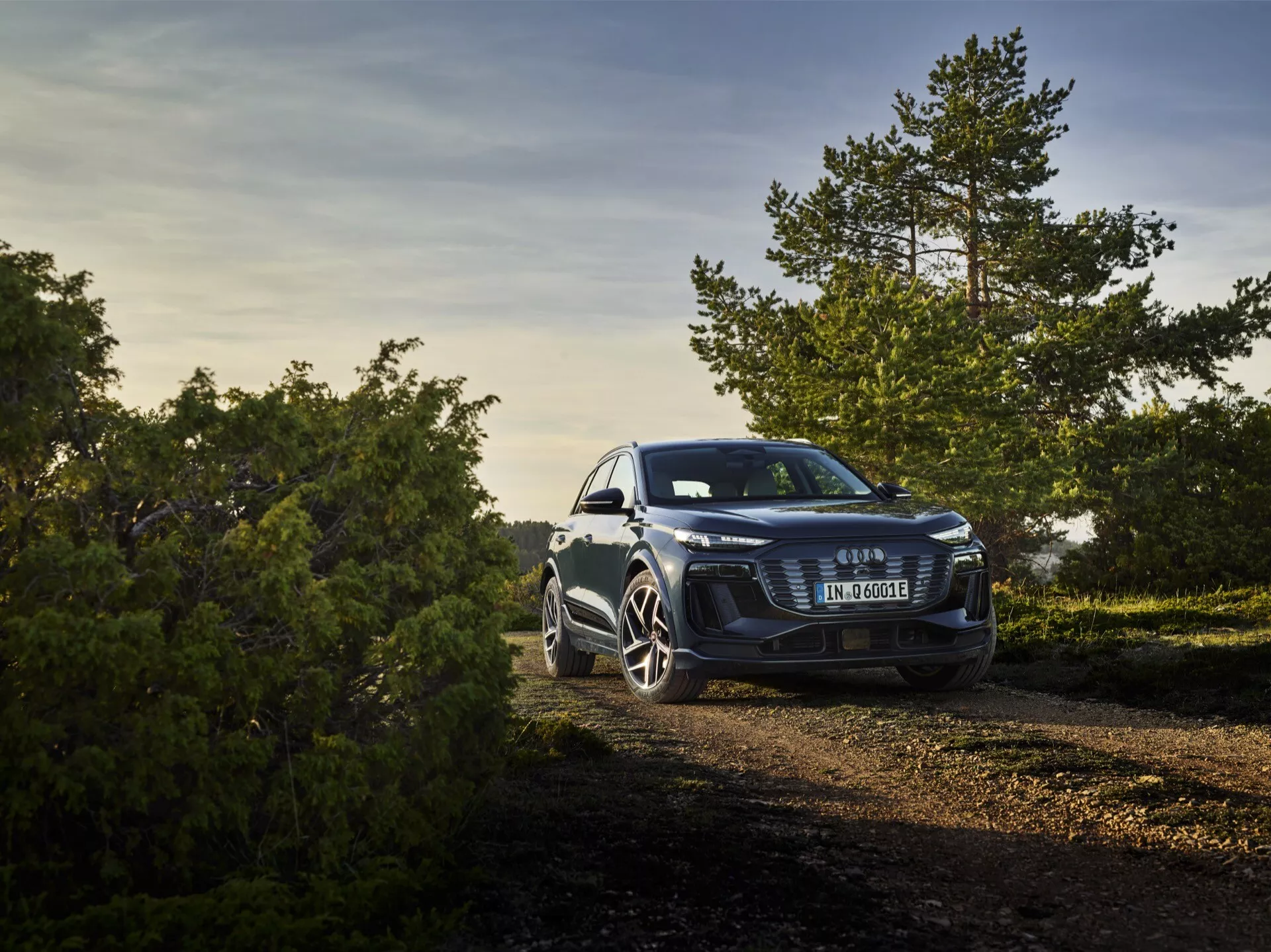 Vue avant trois-quarts de l'Audi Q6 e-tron 2024 garée en plein air sur un sentier de terre en pleine nature, prise à la lumière chaude et dorée du début de matinée ou de fin d'après-midi. L'angle surélevé met en valeur la calandre audacieuse avec un emblème centré, des phares élégants suggérant une technologie d'éclairage LED ou Matrix LED avancée et des roues en alliage sportives distinctives. La photo comprend des éléments naturels en arrière-plan, indiquant les capacités polyvalentes de la voiture tant en milieu urbain que pour des activités légères en dehors de la route.