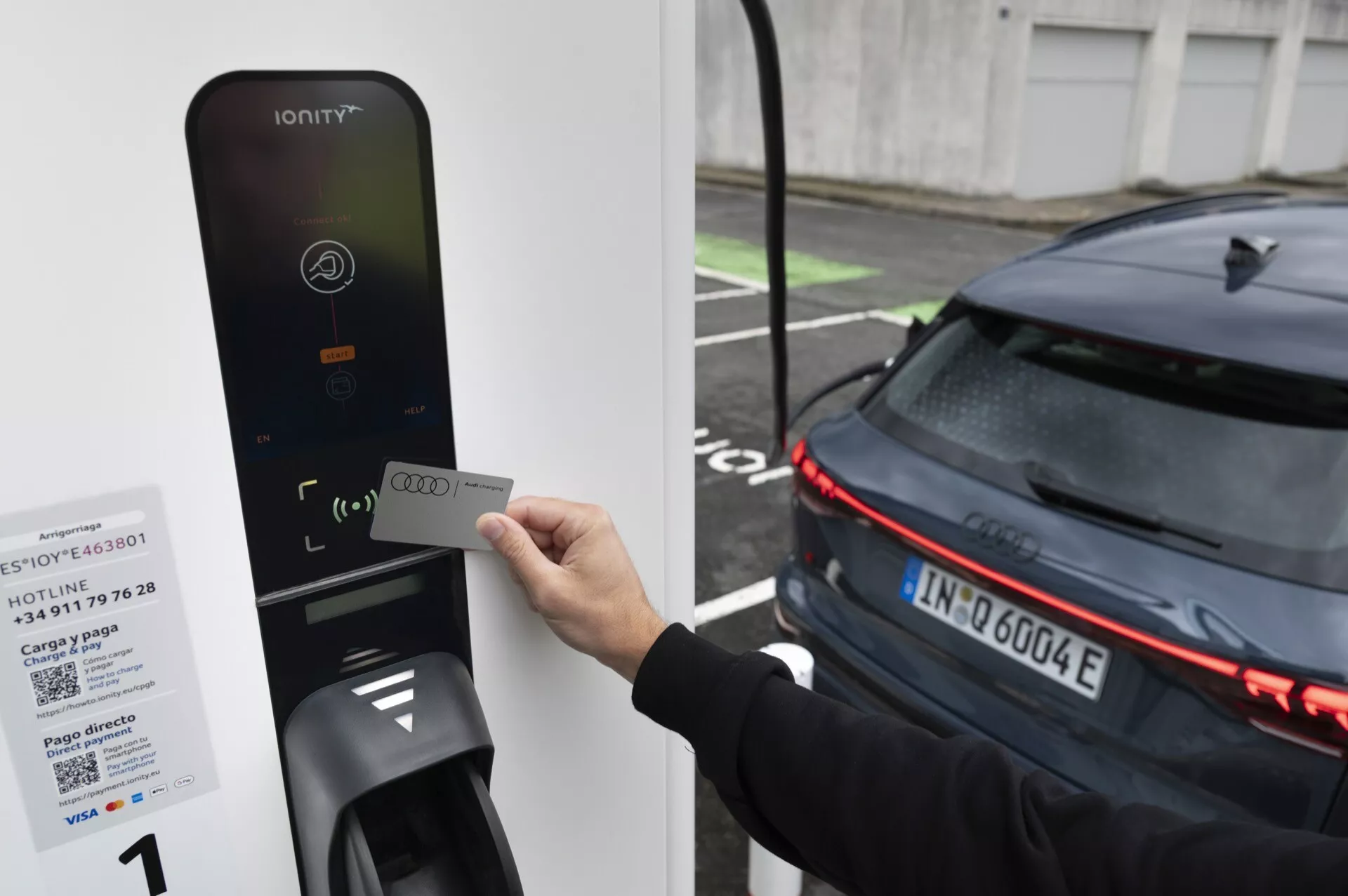 Image d'une personne tenant une carte RFID près du lecteur d'une station de recharge pour véhicules électriques IONITY, dirigée vers l'arrière d'une voiture moderne de couleur sombre avec des feux arrière à LED horizontaux, indiquant sa nature électrique. La voiture, vraisemblablement un VE, est stationnée à côté du chargeur, suggérant qu'elle est en cours de recharge ou sur le point de se recharger.