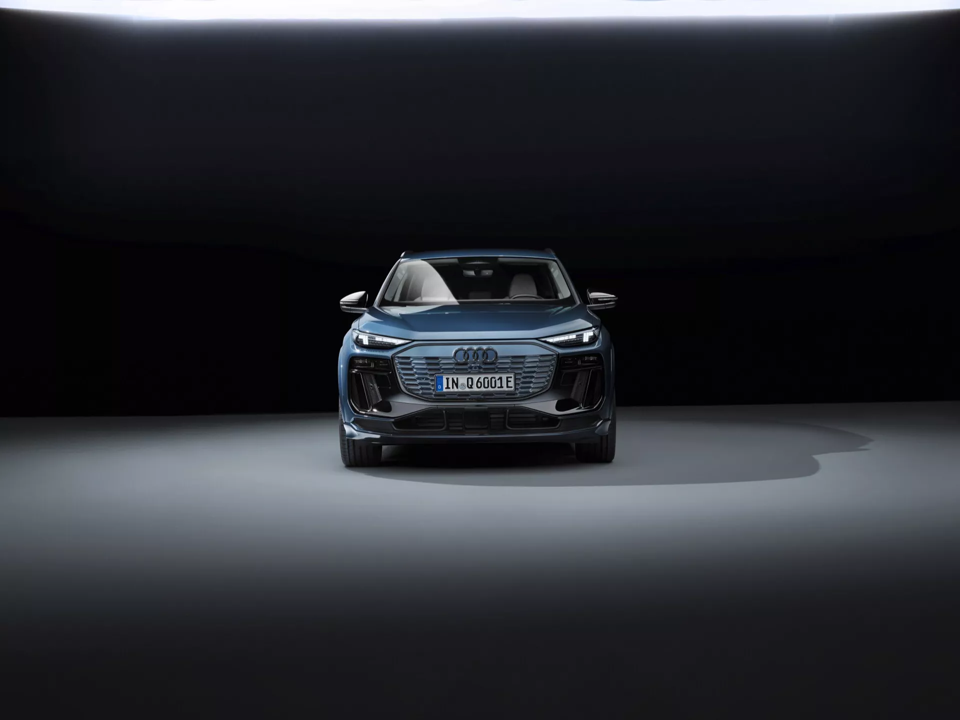Vue avant de l'Audi Q6 e-tron 2024, mettant en avant le capot, la grille, une partie du toit et le design avant. Les phares élégants sont probablement équipés de la technologie LED. La plaque d'immatriculation allemande suggère la nature électrique ou hybride du véhicule. Grille distinctive Audi Singleframe, prises d'air supplémentaires et capteurs de sécurité potentiels, soulignant ses technologies avancées d'assistance à la conduite. L'éclairage dramatique avec un arrière-plan sombre et une illumination par le haut met en valeur le design luxueux et sophistiqué de la voiture.