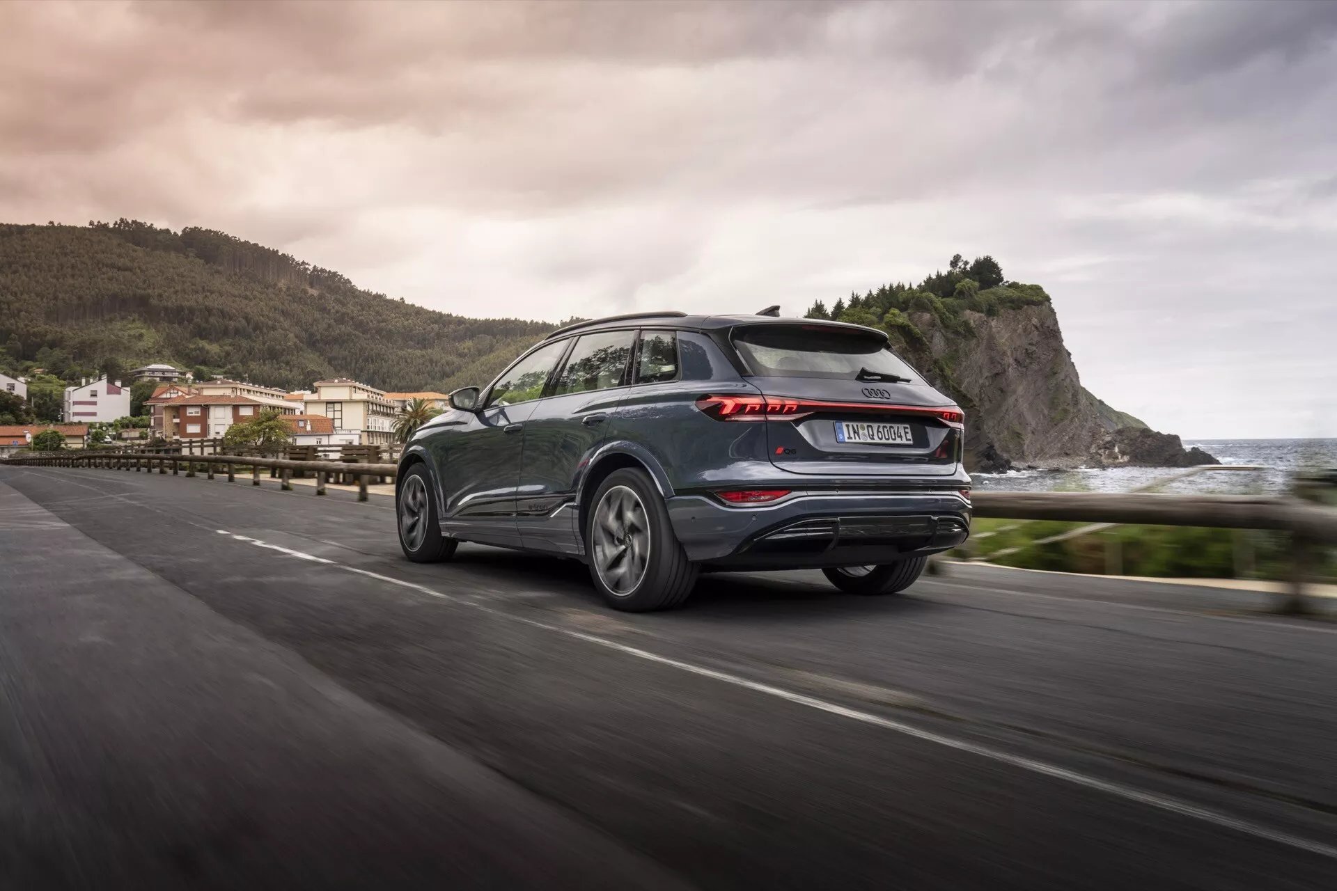 Vue arrière trois quarts de l'Audi Q6 e-tron 2024 en mouvement sur une route côtière, mettant en évidence des feux arrière à LED distinctifs, une couleur métallique sombre, des jantes en alliage et un essuie-glace arrière pratique. L'arrière-plan flou suggère un environnement côtier, reflétant les avancées esthétiques et technologiques d'un SUV moderne.