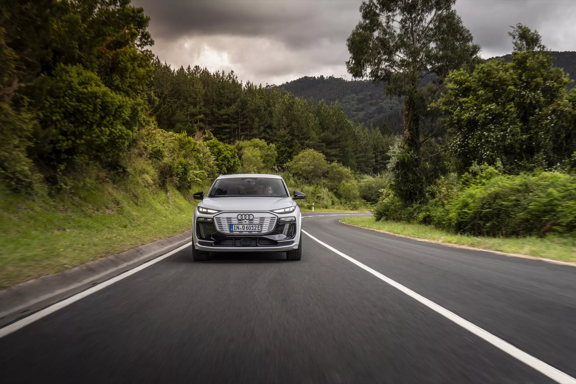 Audi Q6-e-tron 2024 capturée en mouvement depuis un angle frontal sur une route à deux voies bordée de verdure. La photo montre la calandre, les phares, le capot et le pare-chocs avant du véhicule, mettant en avant ses performances dynamiques et son design. La voiture dispose de phares LED distinctifs, d'un emblème de luxe proéminent sur la calandre et de lignes aérodynamiques, le tout sous un temps nuageux avec des conditions de route sèche.