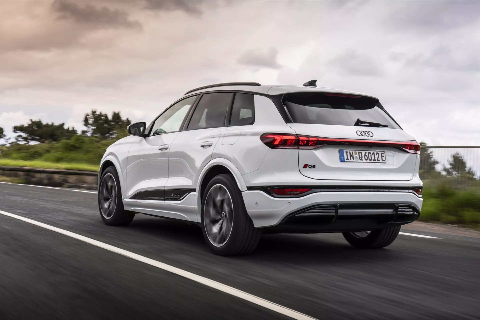 Audi Q4 e-tron 2024 en mouvement sur une route goudronnée vue en trois quarts arrière, capturant le côté arrière gauche. L'image montre des feux arrière fins, un becquet de toit, un toit panoramique, des jantes en alliage à plusieurs rayons, une carrosserie bicolore avec garniture métallique, et l’emblème emblématique des anneaux Audi. Le flou de mouvement des roues et la verdure en bord de route avec un ciel couvert suggèrent des conditions de conduite dynamiques et par tous les temps.