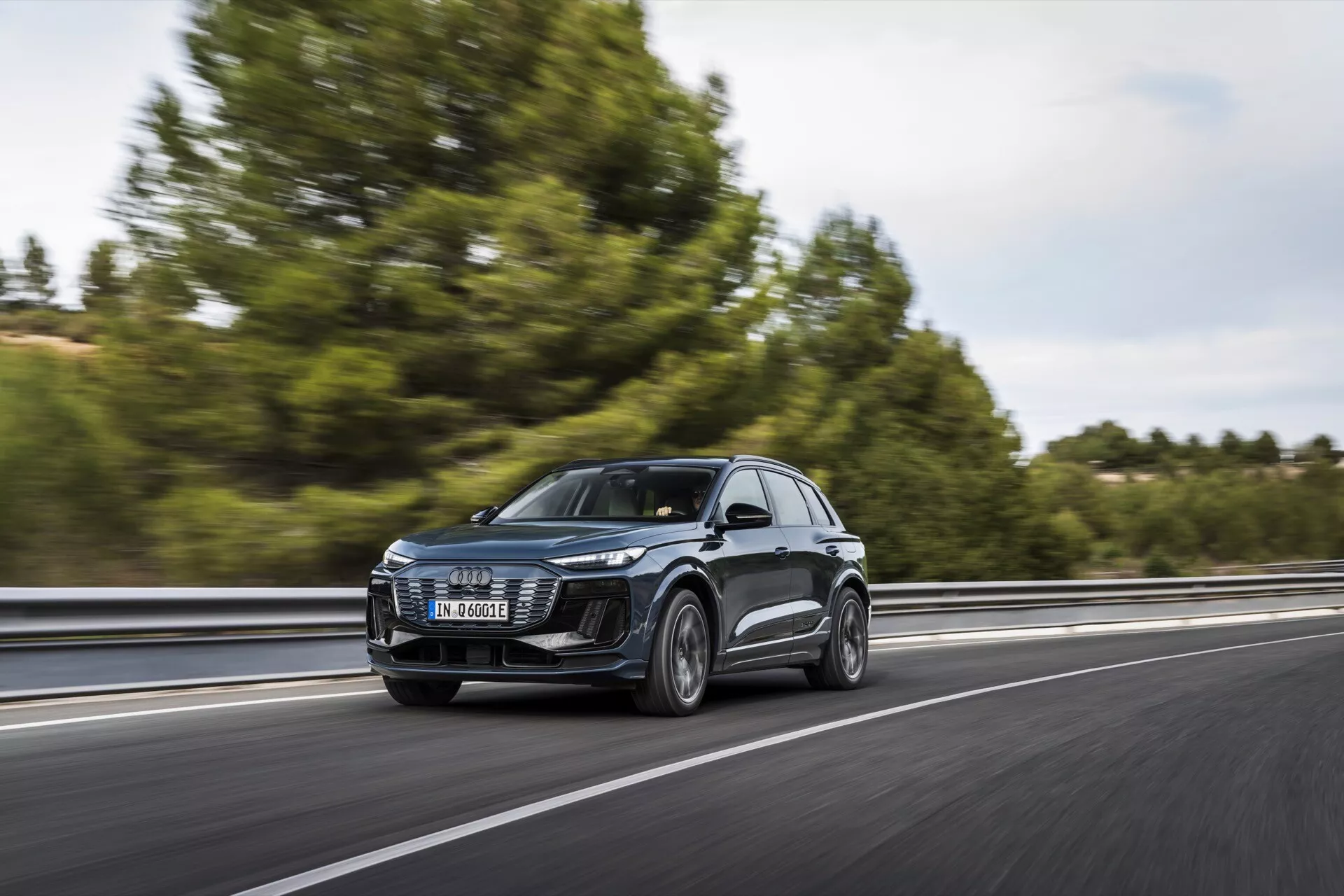 L'Audi Q6 e-tron 2024 SUV en mouvement sur une autoroute, capturé depuis une vue avant trois quarts. L'image montre la calandre avant, les phares à LED, le capot, le pare-brise et une partie du profil latéral, y compris les portes et les rétroviseurs latéraux. Le véhicule roule sur le côté droit de la voie, se déplaçant de gauche à droite avec un fond flou indiquant la vitesse. Les arbres et un garde-corps sont légèrement visibles. Le design clair des phares avant suggère des systèmes d'éclairage avancés et la grande calandre suggère une technologie de refroidissement ou des capteurs pour l'ADAS. Les rétroviseurs latéraux peuvent abriter des indicateurs de direction ou des systèmes de surveillance des angles morts. Les roues donnent un aperçu des catégories de performances ou de luxe.
