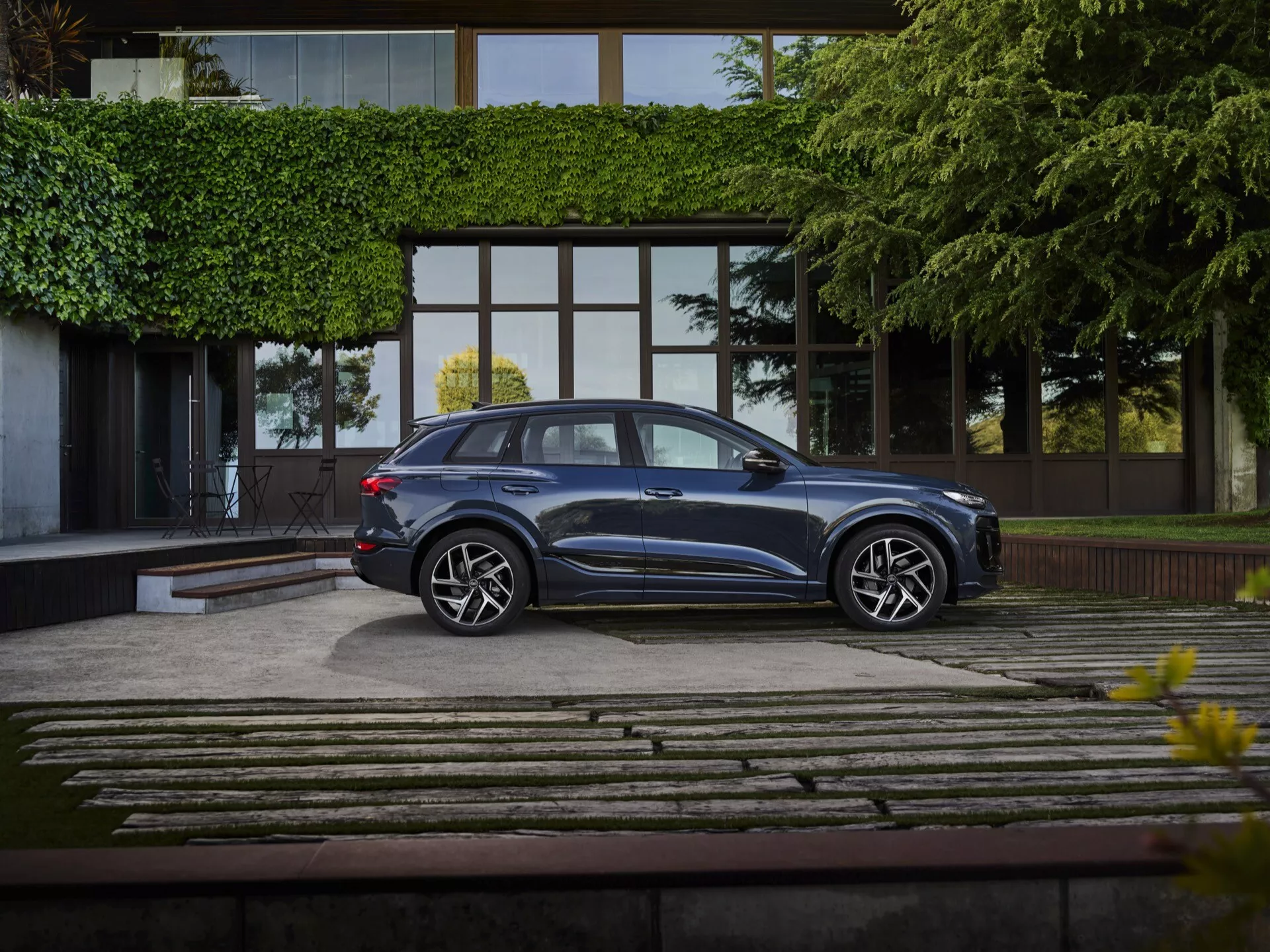Une Audi Q6 e-tron 2024 est garée dans une allée bien aménagée, capturée en vue de profil complet sous un angle légèrement élevé. Le SUV bleu métallique profond brille avec un design élégant, présentant des lignes de toit aérodynamiques, des fenêtres teintées et des poignées de porte affleurantes. Les roues en alliage multibrins et les pneus à profil bas soulignent sa finition haut de gamme. Positionnée devant un bâtiment moderne et luxueux avec de grandes fenêtres en verre et une allée de pierre et d'herbe, la scène est doucement éclairée, mettant en valeur le statut premium du véhicule.