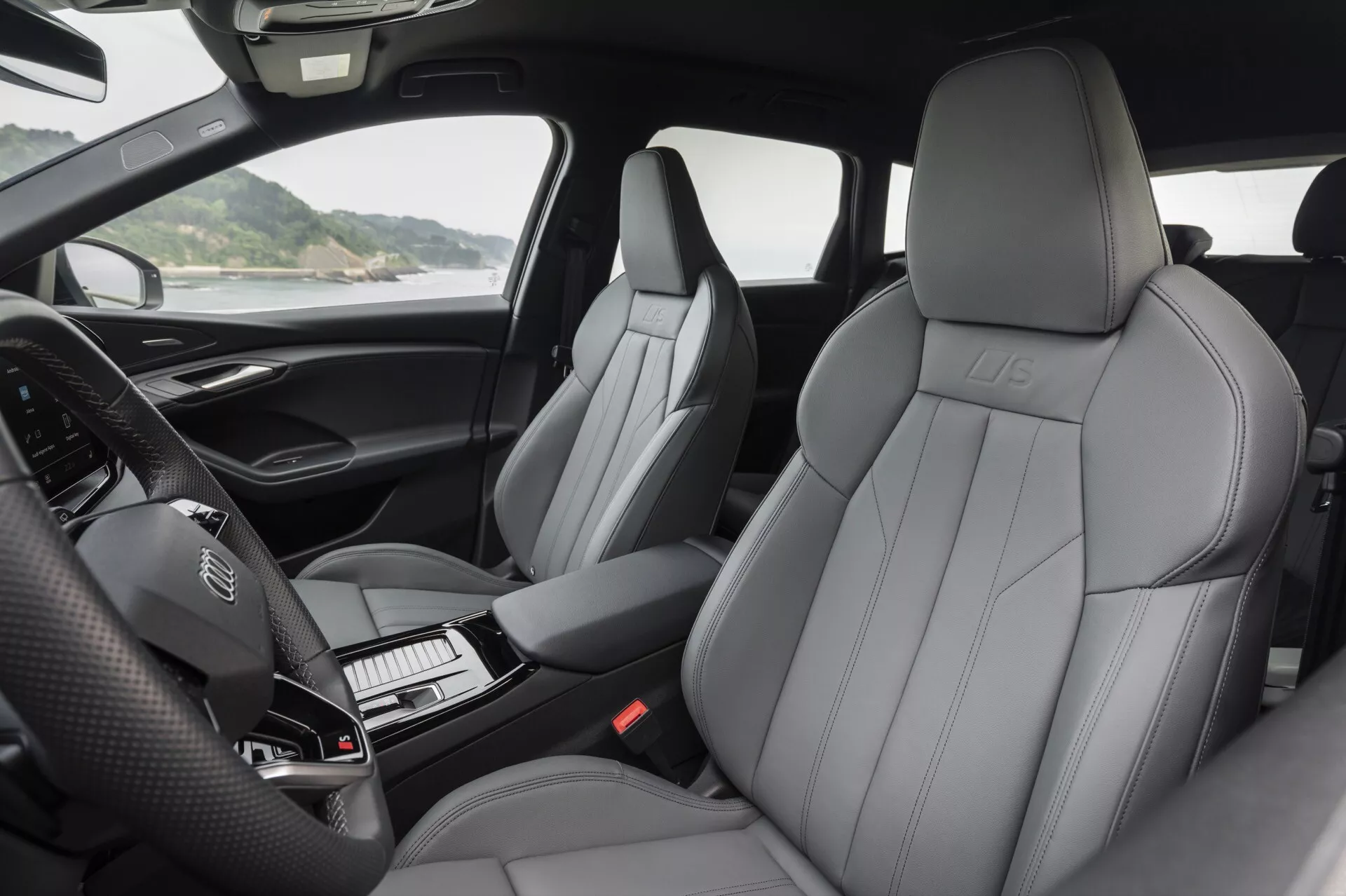Vue intérieure de l'Audi Q6 e-Tron 2024 présentant des sièges avant en cuir luxueux avec un motif de couture prononcé, un logo embossé et un design contemporain. La photographie met en avant le tableau de bord avec un volant à commandes intégrées et un combiné d'instruments numérique éteint. La console centrale qui abrite le levier de vitesses et des boutons ordonnés est partiellement visible. L'image capture les accoudoirs des portes capitonnés avec des commandes pour les rétroviseurs, les fenêtres et possiblement les sièges. La lumière naturelle accentue le confort et la haute qualité des matériaux de l'habitacle.