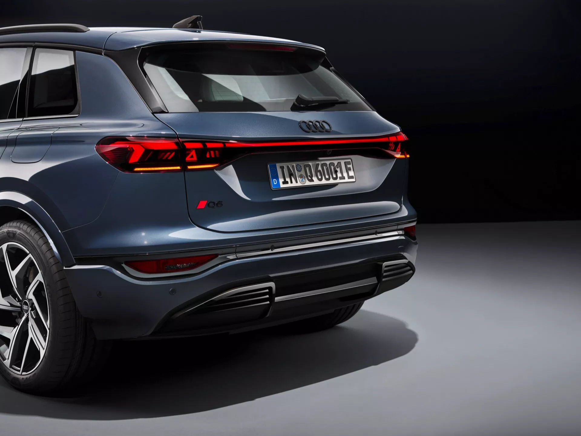 Cette image montre un SUV moderne, probablement l'Audi Q6 e-tron 2024, vu sous un angle arrière élevé. L'emblématique emblème Audi à quatre anneaux est bien en évidence à l'arrière, entouré d'une élégante bande de feux arrière à LED continue qui s'étend sur toute la largeur du véhicule, avec des feux de freinage et des clignotants segmentés. La voiture bleu marine foncé présente une finition métallique polie, un design audacieux du pare-chocs avec échappements intégrés et diffuseur, des jantes en alliage multirayons avec des pneus à profil bas, et une plaque d'immatriculation suggérant une motorisation électrique ou hybride. L'éclairage professionnel et le fond neutre mettent en avant le design luxueux et avancé du véhicule, soulignant ses attributs sportifs et haut de gamme.