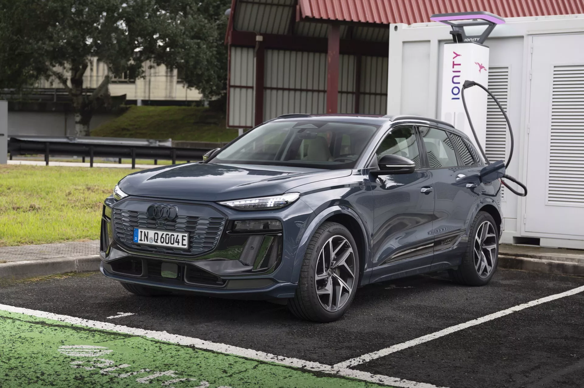 L'image montre un SUV moderne Audi Q6 e-Tron 2024 garé à une station de recharge pour véhicules électriques. La voiture est présentée sous un angle légèrement frontal, mettant en évidence sa calandre, ses phares et une partie du capot et du pare-brise. Elle est positionnée en diagonale avec l'avant orienté légèrement à gauche. Le côté gauche du véhicule, incluant la roue avant côté conducteur, la porte et le rétroviseur, est visible. Le SUV est branché à une station de recharge sur le côté avant gauche au dessus de la roue. La voiture présente des lignes élégantes, une calandre fermée typique des véhicules électriques et des phares LED avancés. Les détails suggèrent la présence de systèmes avancés d'assistance à la conduite comme le régulateur de vitesse adaptatif, l'assistant de maintien de voie et le système d'évitement des collisions. L'image est lumineuse et claire, prise en plein jour sans flou de mouvement, rendant tous les éléments de design et technologiques facilement visibles.