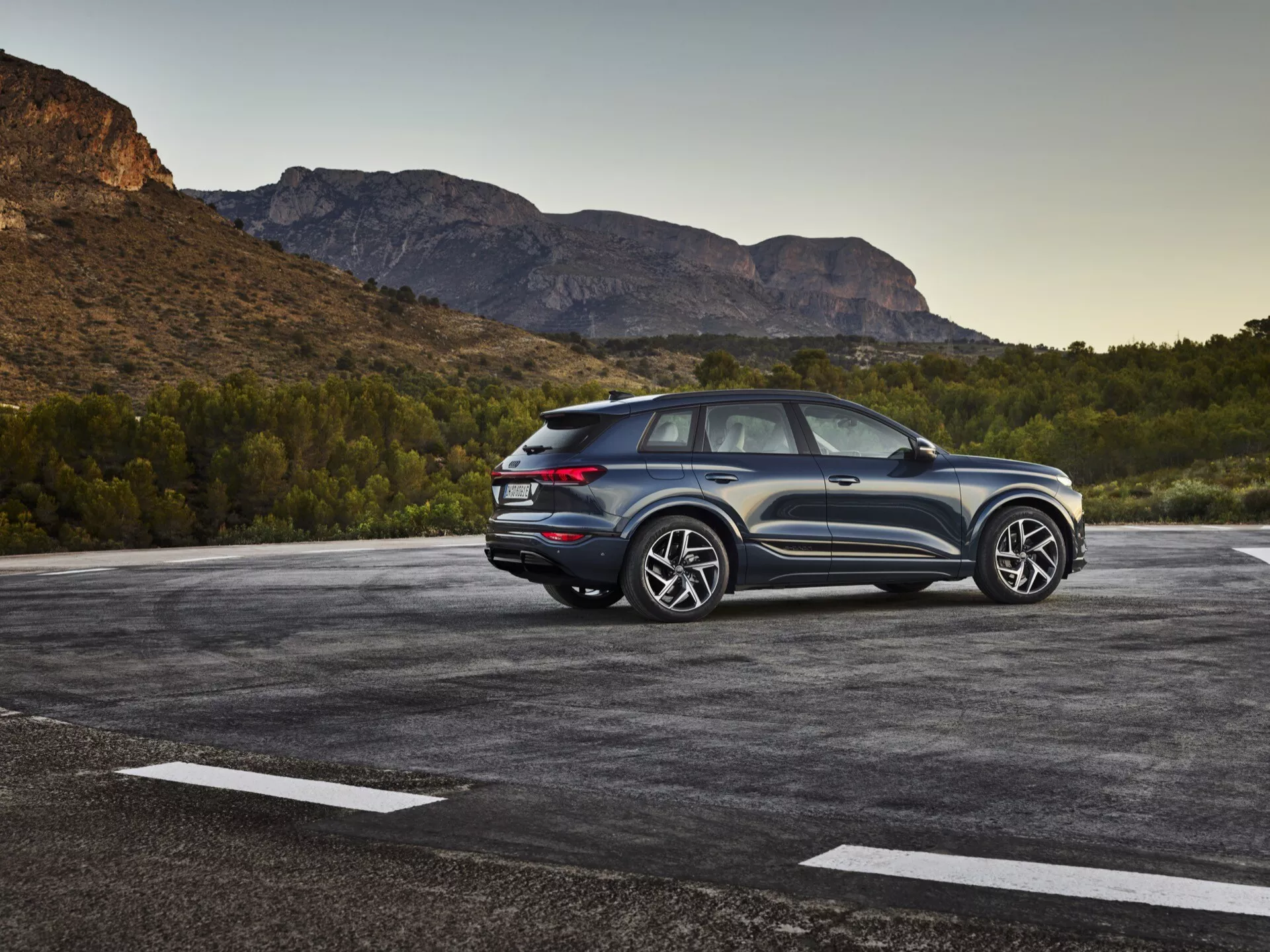 Image d'un SUV Audi Q6-e-tron 2024 garé sur un parking extérieur avec un paysage naturel, peut-être montagneux, en arrière-plan. La photo capture une vue arrière aux trois quarts, mettant en valeur les éléments de design latéraux et arrière. Le côté conducteur est clairement visible, montrant les lignes de design, les fenêtres arrière et le design des roues. La ligne de toit inclinée suggère un design de SUV coupé sportif. Les feux arrière à LED modernes, les capteurs ou réfléchisseurs visibles sur le pare-chocs arrière et le schéma de couleurs bicolore accentuent la hauteur et l'allure. Les grandes roues en alliage et les pneus substantiels indiquent un niveau de finition supérieur avec une esthétique sportive. Le véhicule semble proche de l'appareil photo, offrant une vue détaillée. Les caractéristiques notables incluent l'éclairage LED, les capteurs de stationnement, les roues et pneus haute performance, le design aérodynamique, et possiblement les systèmes avancés d'assistance à la conduite et la traction intégrale.