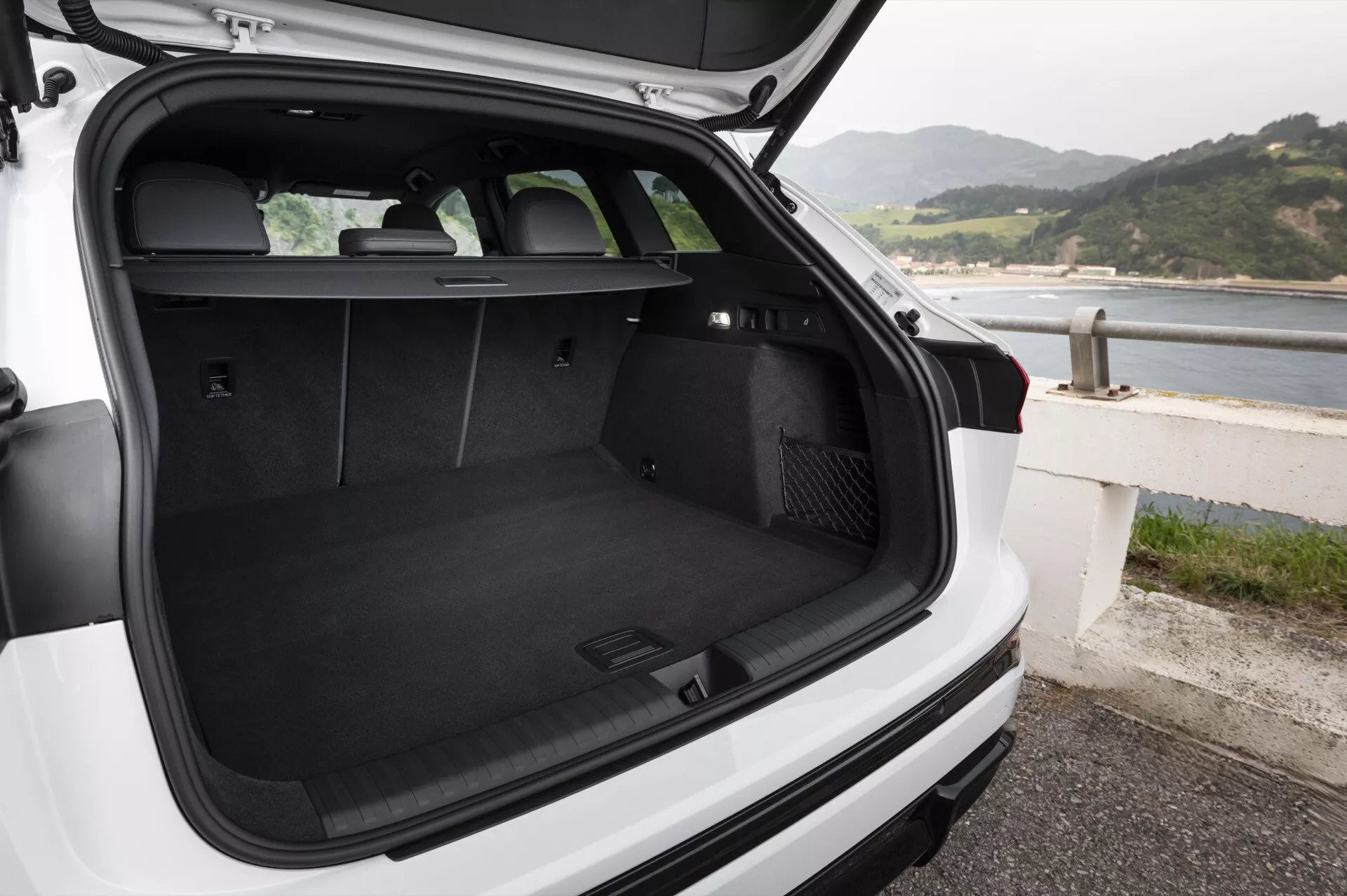 L'image montre l'espace de chargement d'une Audi Q6 e-Tron 2024 blanche de type hatchback ou SUV, vue de l'arrière avec le hayon ouvert et un angle de caméra légèrement vers le bas. Le coffre tapissé de noir est vide, démontrant son ample espace et son état impeccable. Les sièges arrière sont en position verticale, indiquant la possibilité de transporter des passagers ou de rabattre les sièges pour plus d'espace de chargement. Un cache-bagages rétractable est positionné juste derrière les sièges arrière mais n'est pas étendu, permettant une vue complète du coffre. Sur le côté gauche de l'espace de chargement se trouve une prise de courant de 12 volts, et du côté droit semble se trouver un compartiment probablement destiné à ranger de petits objets. L'essuie-glace arrière est visible. Le véhicule est garé près d'une côte, suggérant un emplacement en bord de mer. Il n'y a ni objets, ni personnes dans le coffre.