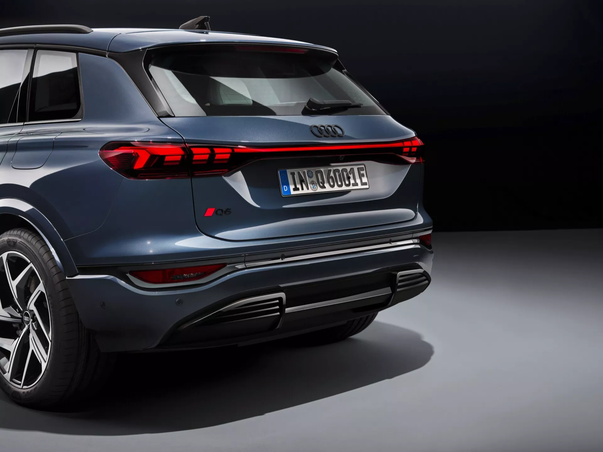 Angle arrière du SUV Audi Q6 e-tron 2024, avec une peinture bleue métallique et des feux arrière LED modernes. L'image met en valeur le design élégant du véhicule avec une prise de vue en contre-plongée mettant en évidence le pare-chocs arrière, les finisseurs d'échappement intégrés, le diffuseur et les grandes jantes en alliage. La signature lumineuse horizontale distinctive et un spoiler de toit proéminent soulignent son esthétique sportive. La plaque d'immatriculation européenne suggère une motorisation électrique ou hybride, avec un motif de bande de roulement visible indiquant des pneus de performance. Capturé dans un studio avec un éclairage contrôlé.