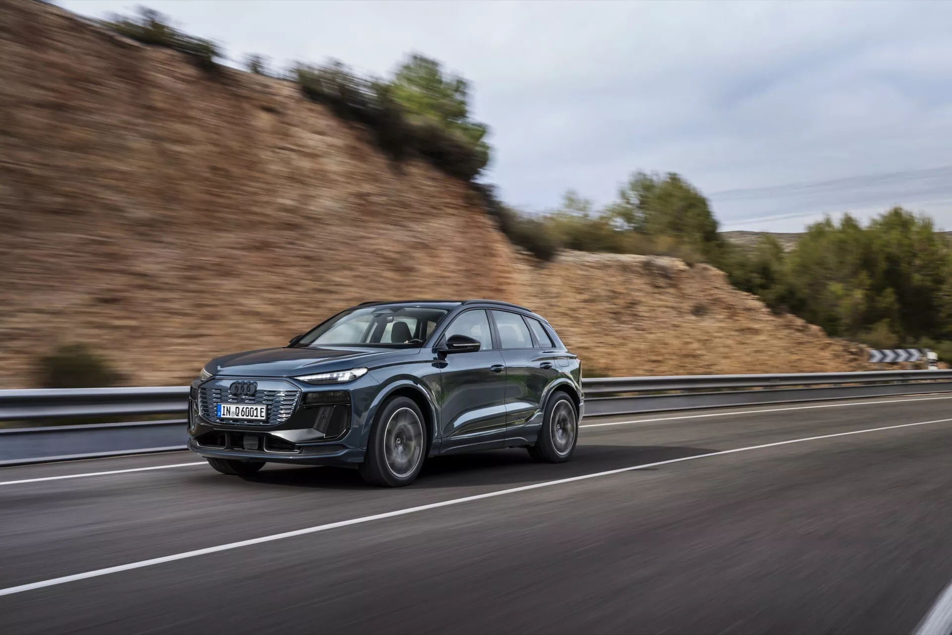 Cette image présente un SUV Audi Q6 e-tron 2024 élégant et sophistiqué, capturé sous un angle latéral en mouvement. Le design moderne du véhicule est souligné par ses phares LED remarquables et ses caractéristiques aérodynamiques, notamment une ligne de toit inclinée et des rétroviseurs latéraux aérodynamiques. La voiture, de couleur sombre, probablement grise ou noire, roule sur une route à grande vitesse, comme en témoigne l'arrière-plan flou créé grâce à une technique photographique de panoramique. La roue avant est légèrement tournée vers l'appareil photo, montrant le design des jantes en alliage. Les autres éléments visibles comprennent une possible technologie intégrée dans les phares et les rétroviseurs latéraux, ainsi que des barres de toit potentiellement axées sur la fonctionnalité.