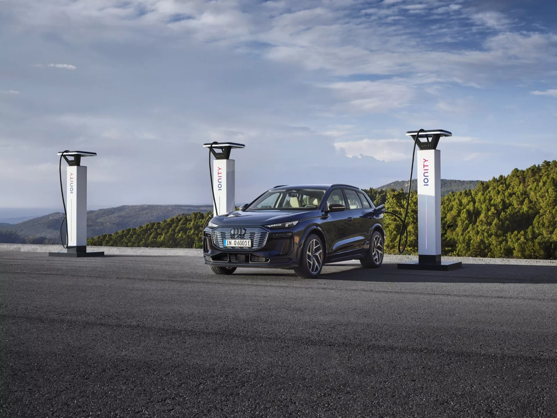 SUV Audi Q6 e-tron 2024 en bleu métallisé profond à une station de recharge de véhicules électriques avec trois chargeurs. L'image capture le véhicule sous un angle de trois quarts montrant son côté avant gauche, mettant en valeur la calandre, les phares, les roues et le capot. Le véhicule électrique élégant et moderne présente un design sportif et polyvalent avec des phares agressifs et élégants et des jantes en alliage, soulignant sa technologie automobile durable et avancée.