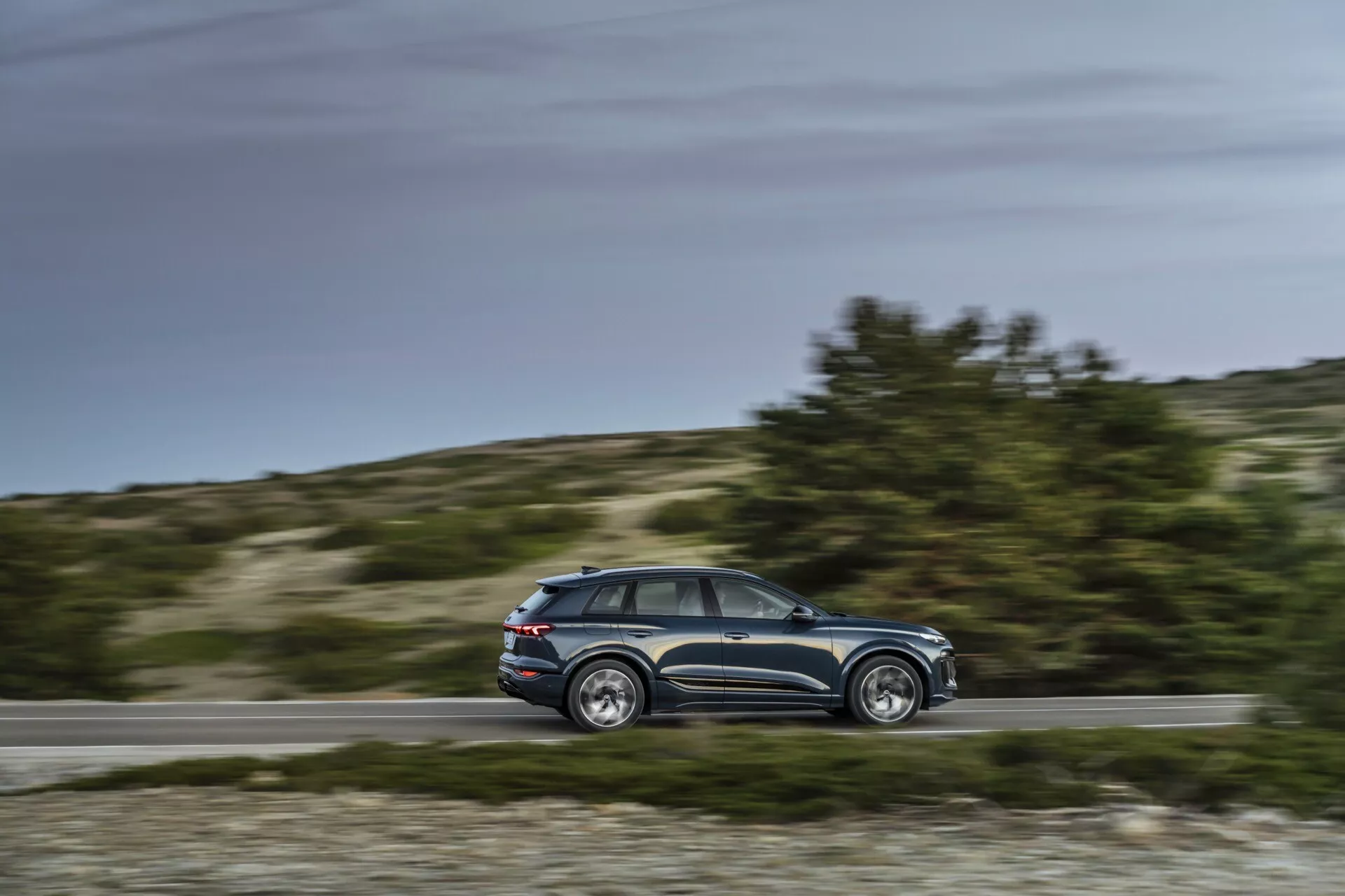 Vue latérale dynamique de l'Audi Q6 e-tron 2024 en mouvement avec fond flou, angle parallèle à distance moyenne montrant le profil, lignes de design, fenêtres, cinq portes, rétroviseurs latéraux, jantes en alliage, configuration diurne, arrière-plan rural.