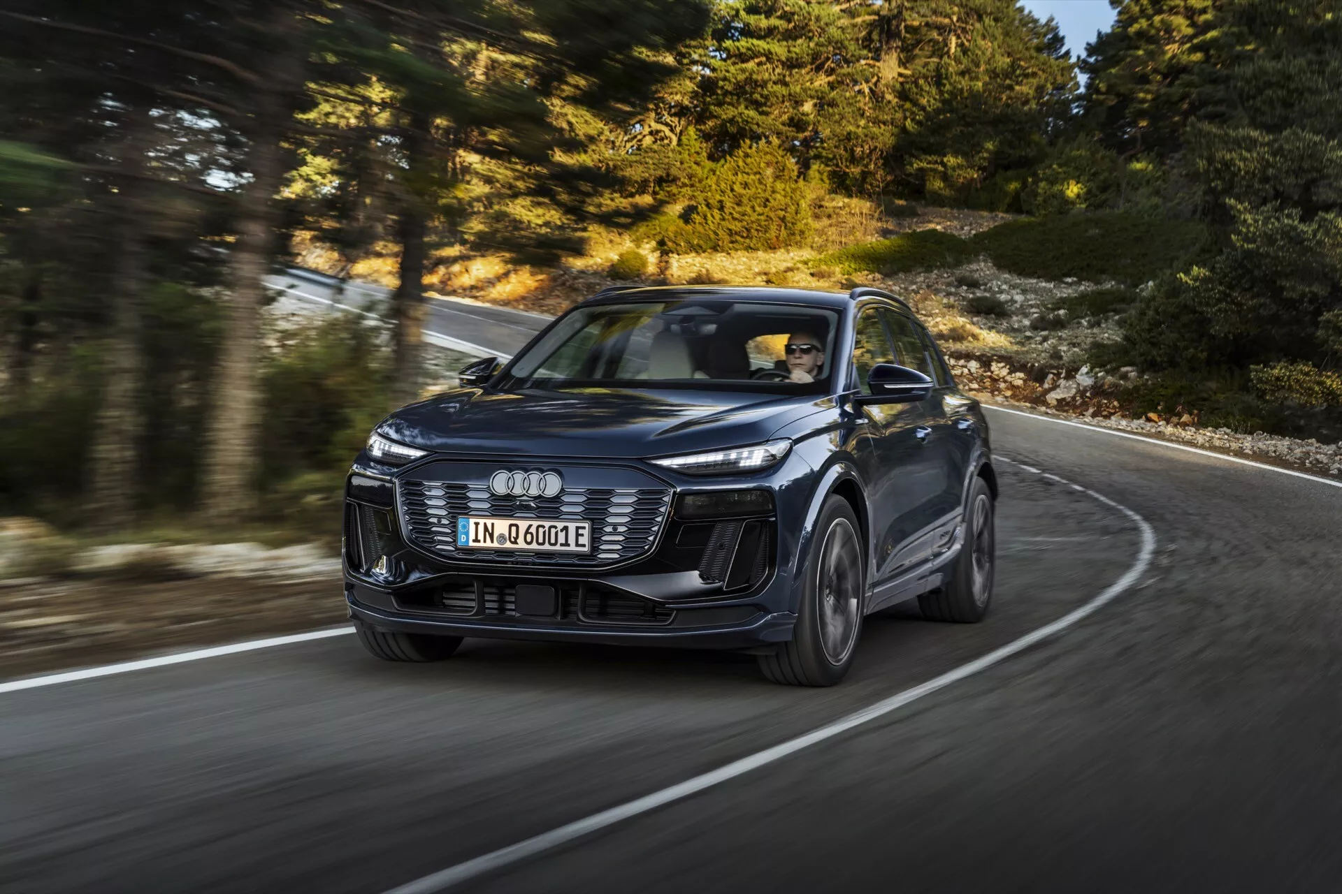 Image d'un SUV Audi Q6 e-tron 2024 moderne en mouvement, capturée sous un angle dynamique de trois quarts avant mettant en lumière son design et ses performances. Le véhicule, identifié par le logo distinctif à quatre anneaux, présente des phares LED emblématiques, un pare-chocs avant agressif et des lignes sculptées sur le capot. L'image, prise sous un angle inférieur, accentue la présence de la voiture sur une route courbée, les roues en alliage étant légèrement floues pour indiquer le mouvement. Le nouveau modèle suggère des technologies avancées, telles que le régulateur de vitesse adaptatif et la conduite électrique, marqué par le badge 'e-tron'. Séance photo professionnelle mettant en avant le luxe, le design et la technologie dans une configuration à conduite à gauche.